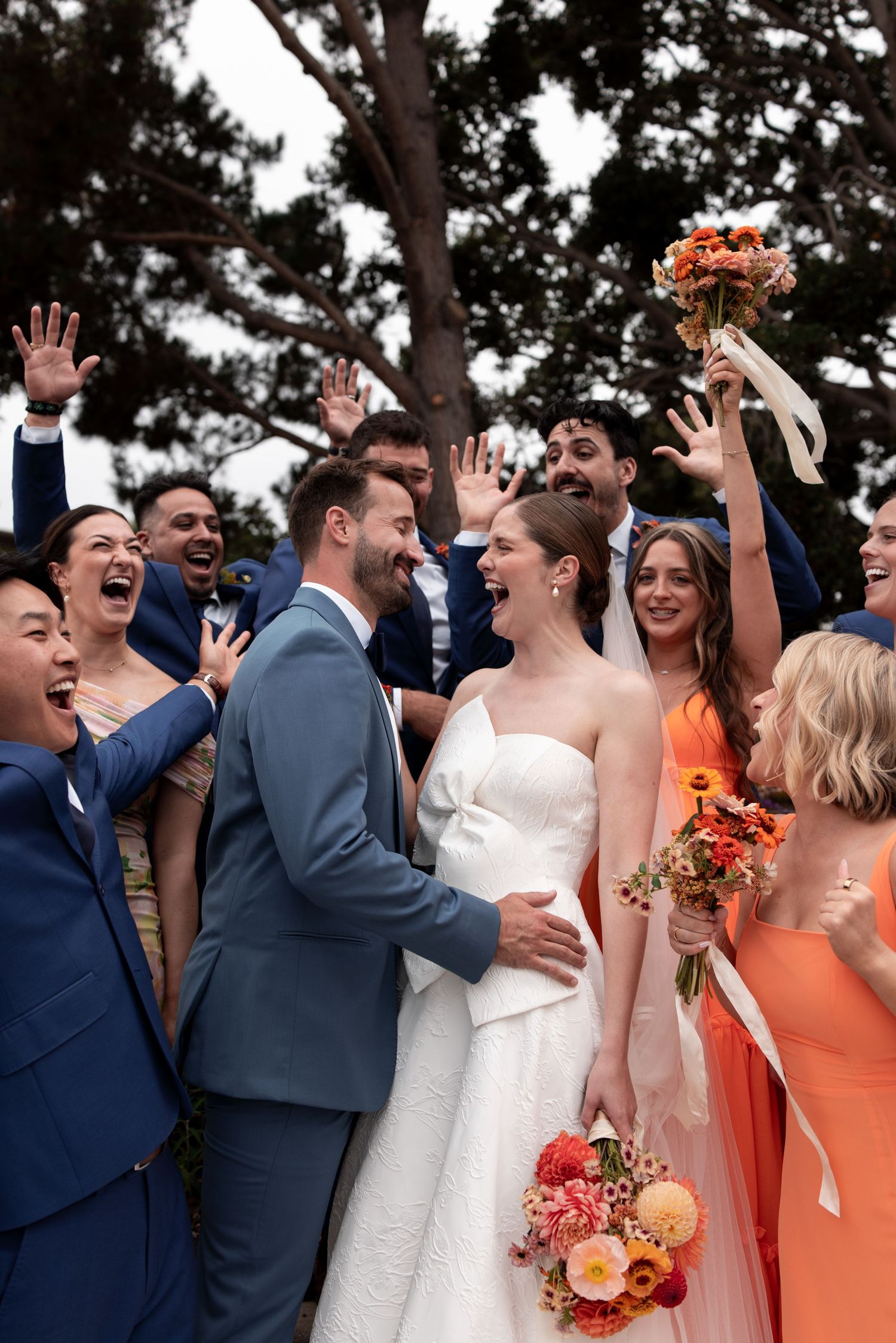 Wedding party laughs together outdoors wearing coral bridesmaid dresses and navy suits at a sunny garden celebration.