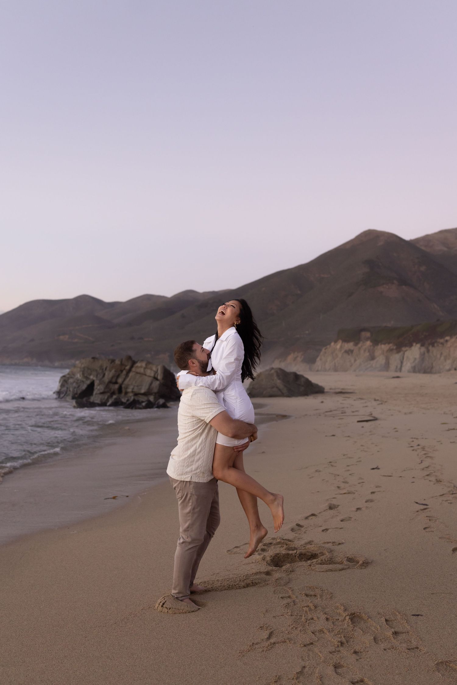 A couple shares a romantic moment on a scenic beach with mountains and cliffs in the background during golden hour.