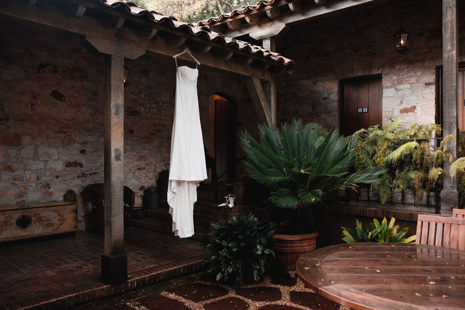 White wedding dress hanging in a rustic stone courtyard with lush tropical plants and wooden beams.
