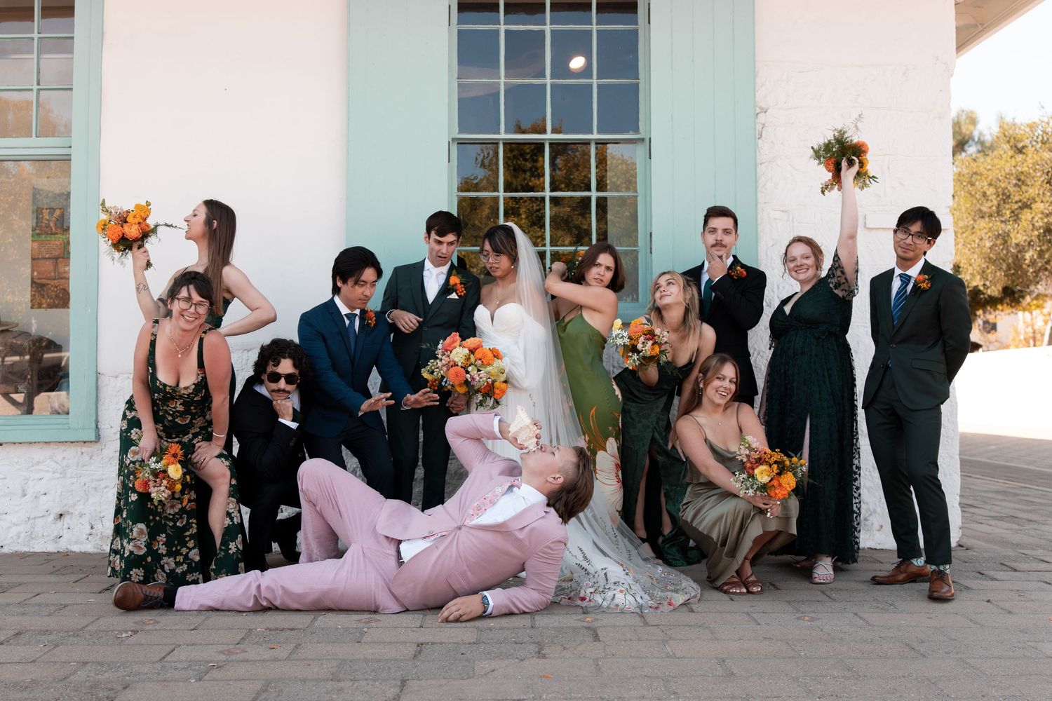 Wedding party members sit together along a white wall in formal attire during outdoor celebration.