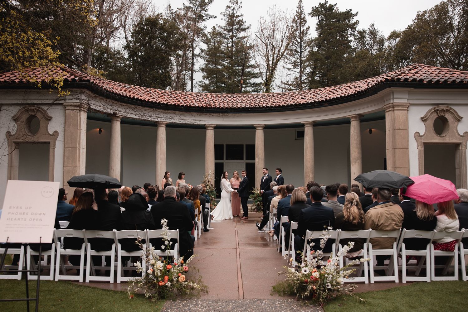 A wedding ceremony takes place in an elegant outdoor courtyard with white columns and Mediterranean-style architecture.