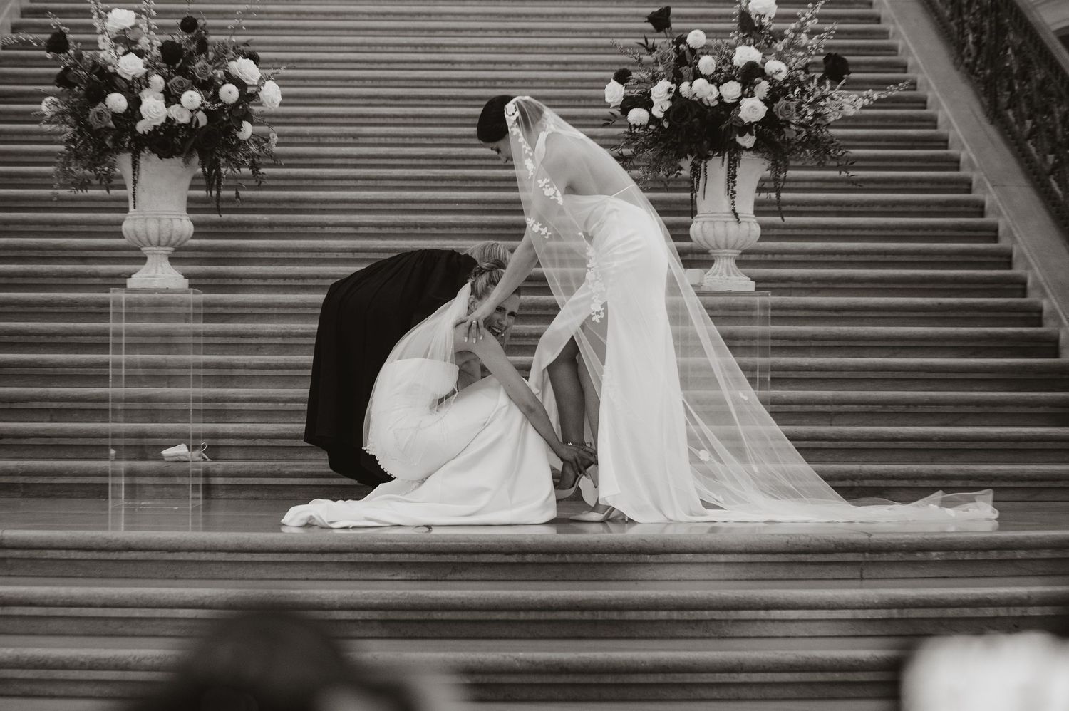 Black and white photo of bride and groom kneeling on grand staircase with floral arrangements during wedding ceremony.