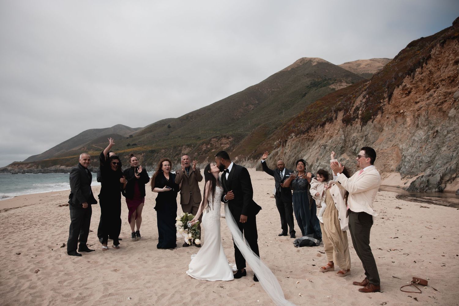 A wedding party gathers along a scenic coastal cliff with dramatic mountains and ocean views in the background.