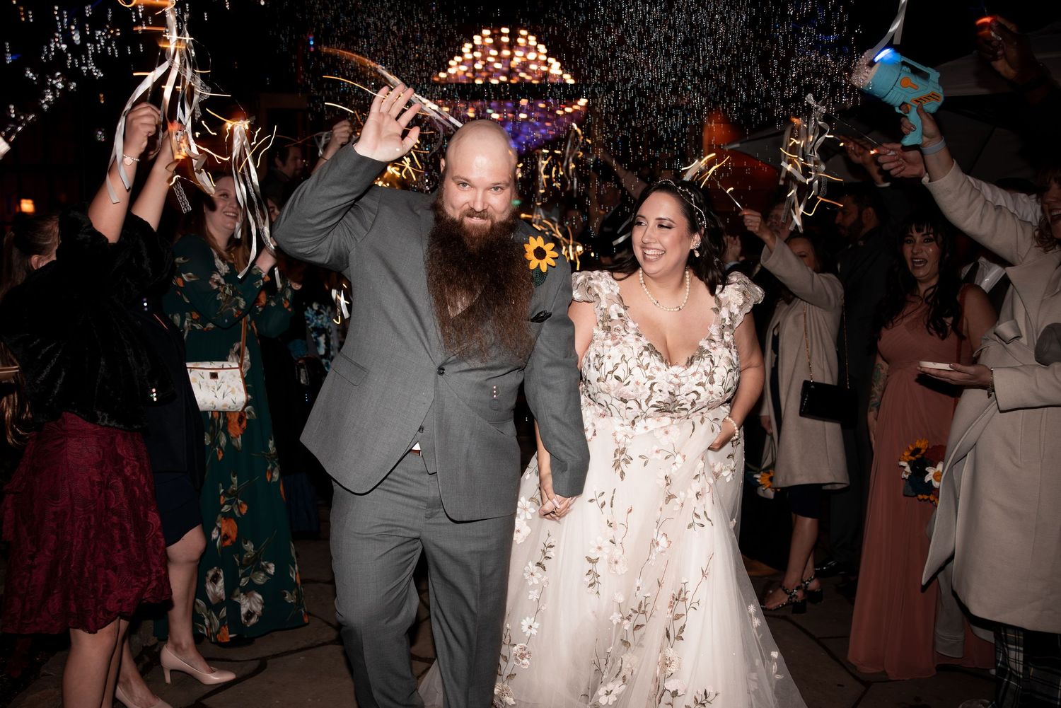 Wedding couple dances joyfully at reception while guests cheer and wave glowsticks in dark venue with sparkling lights.