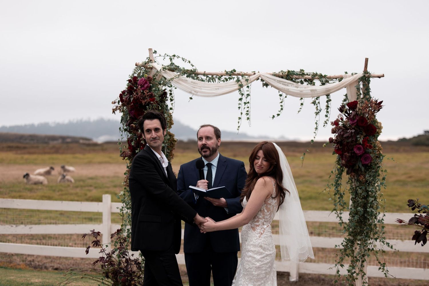 Wedding ceremony participants stand beneath a floral arch in a rustic outdoor setting with mountains in the background.