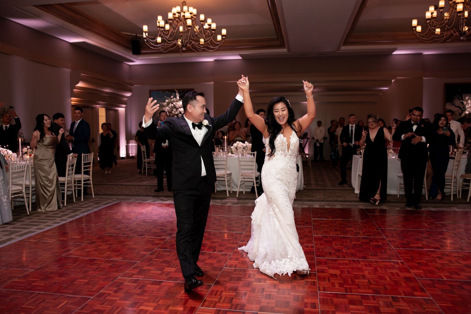 Bride and groom share first dance at elegant wedding reception with chandelier lighting and guests watching.