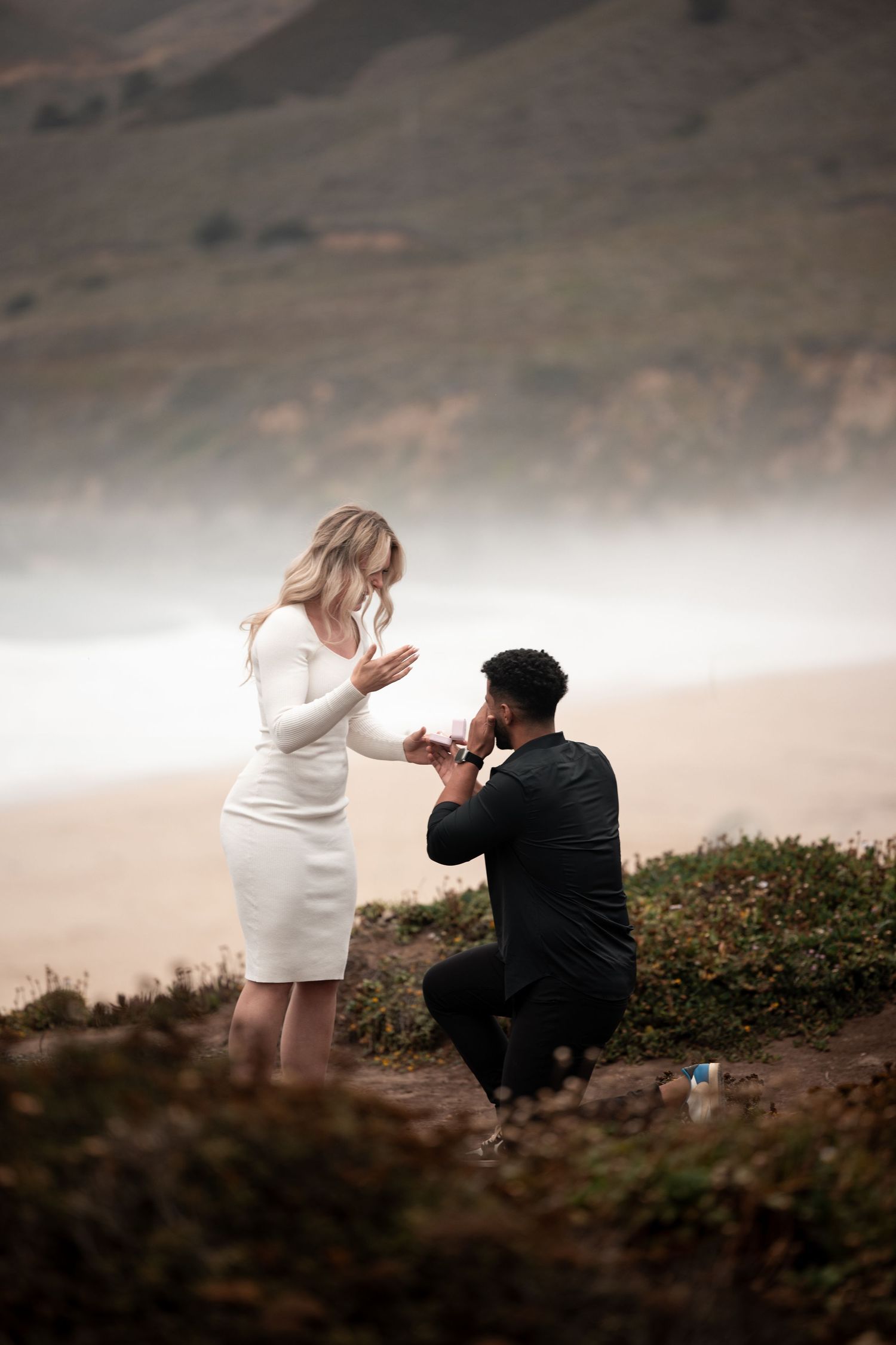 A romantic proposal scene unfolds on a misty coastal cliff with two people in white and black attire.