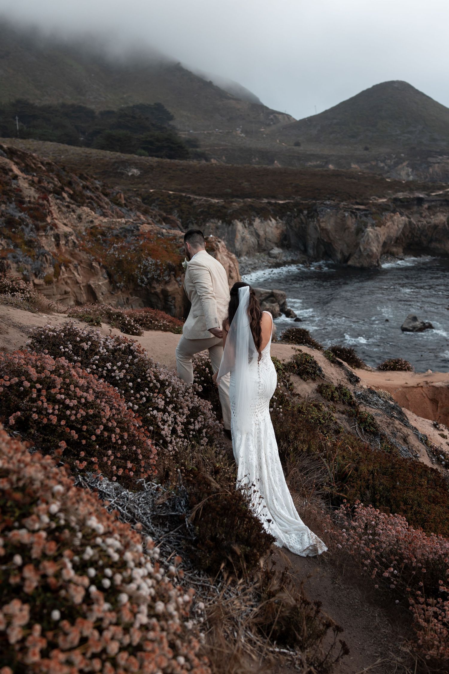 A couple in wedding attire stands on a dramatic coastal cliff overlooking rugged ocean shores on a misty day.