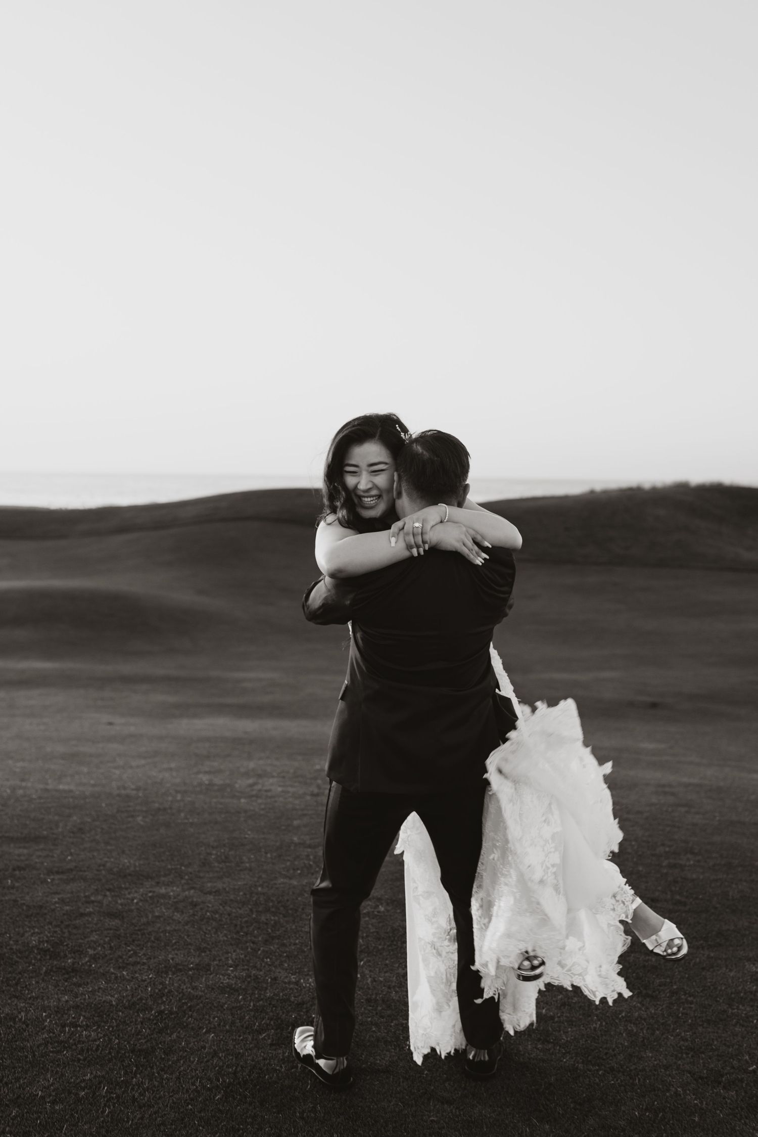Black and white photo of a groom lifting his bride in a wedding dress outdoors on a scenic field.