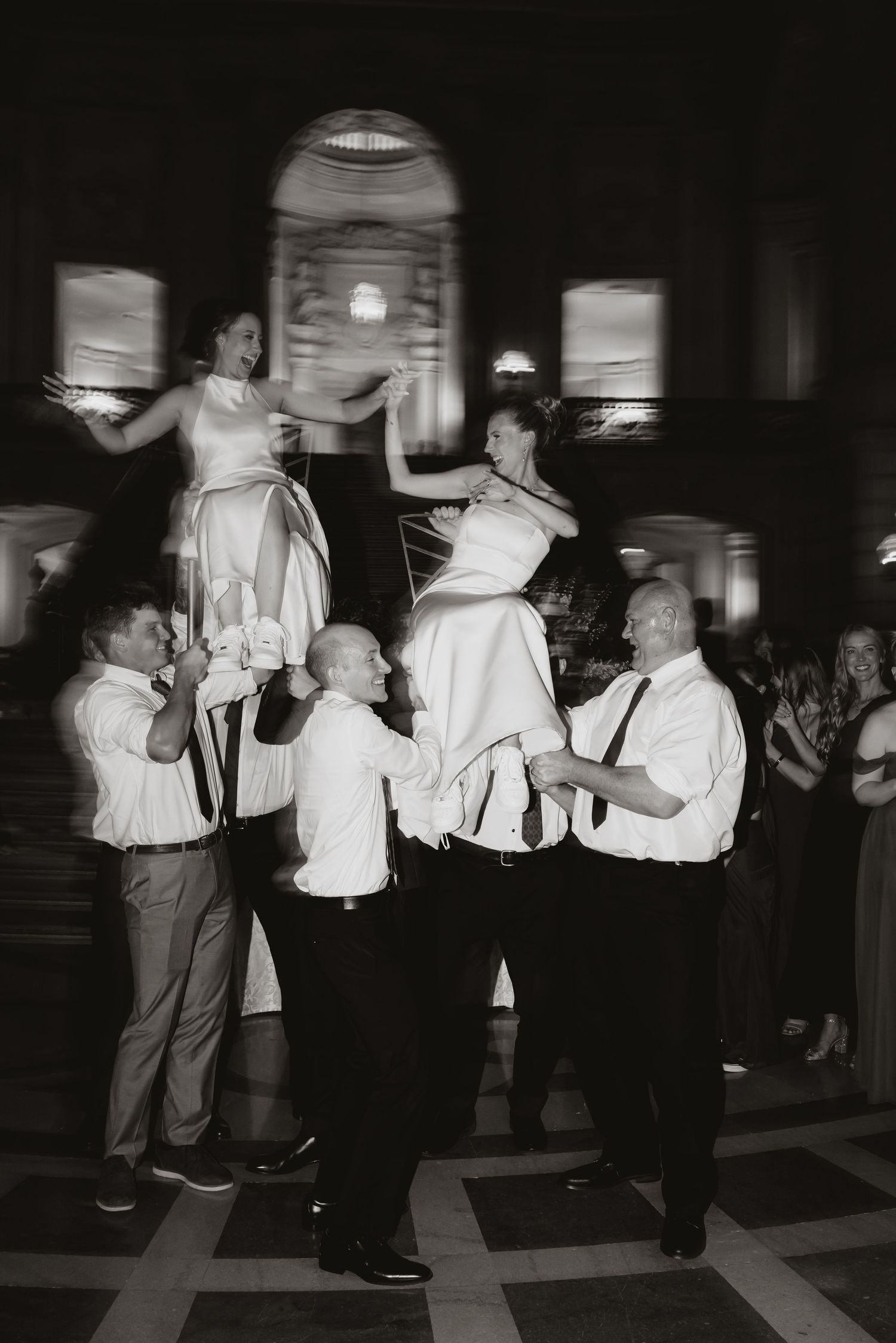 Black and white photo of groomsmen lifting bride and bridesmaid during lively wedding reception dance celebration.