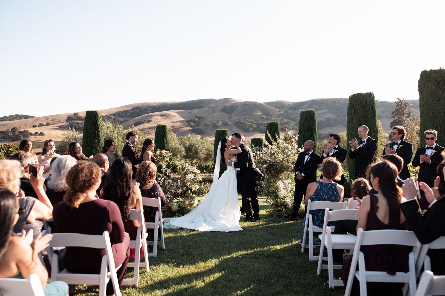 Panoramic view of an outdoor wedding ceremony with white chairs set up on a hillside overlooking scenic mountains.