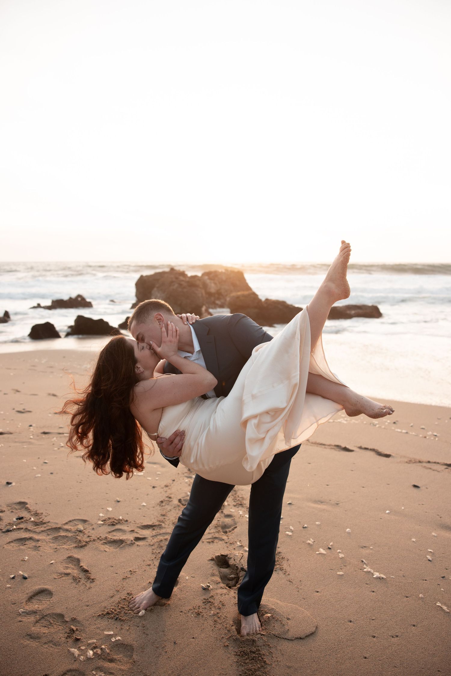 A couple dances playfully together on a sandy beach at sunset, performing romantic dips and spins.