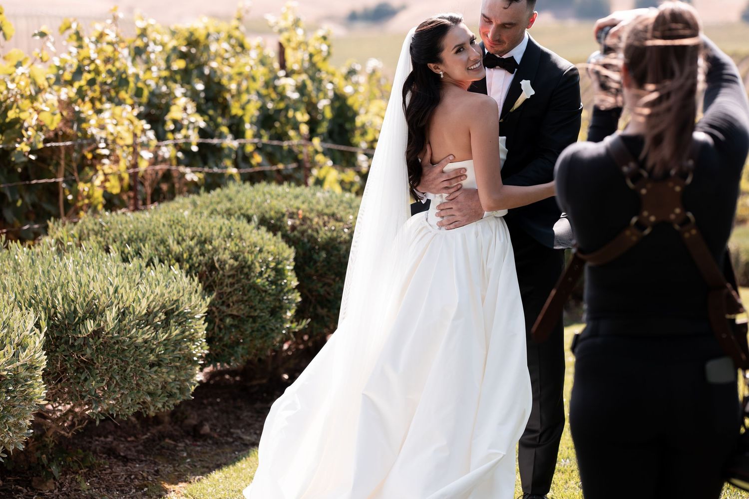 Bride in white gown embraces groom in tuxedo at outdoor vineyard wedding ceremony.