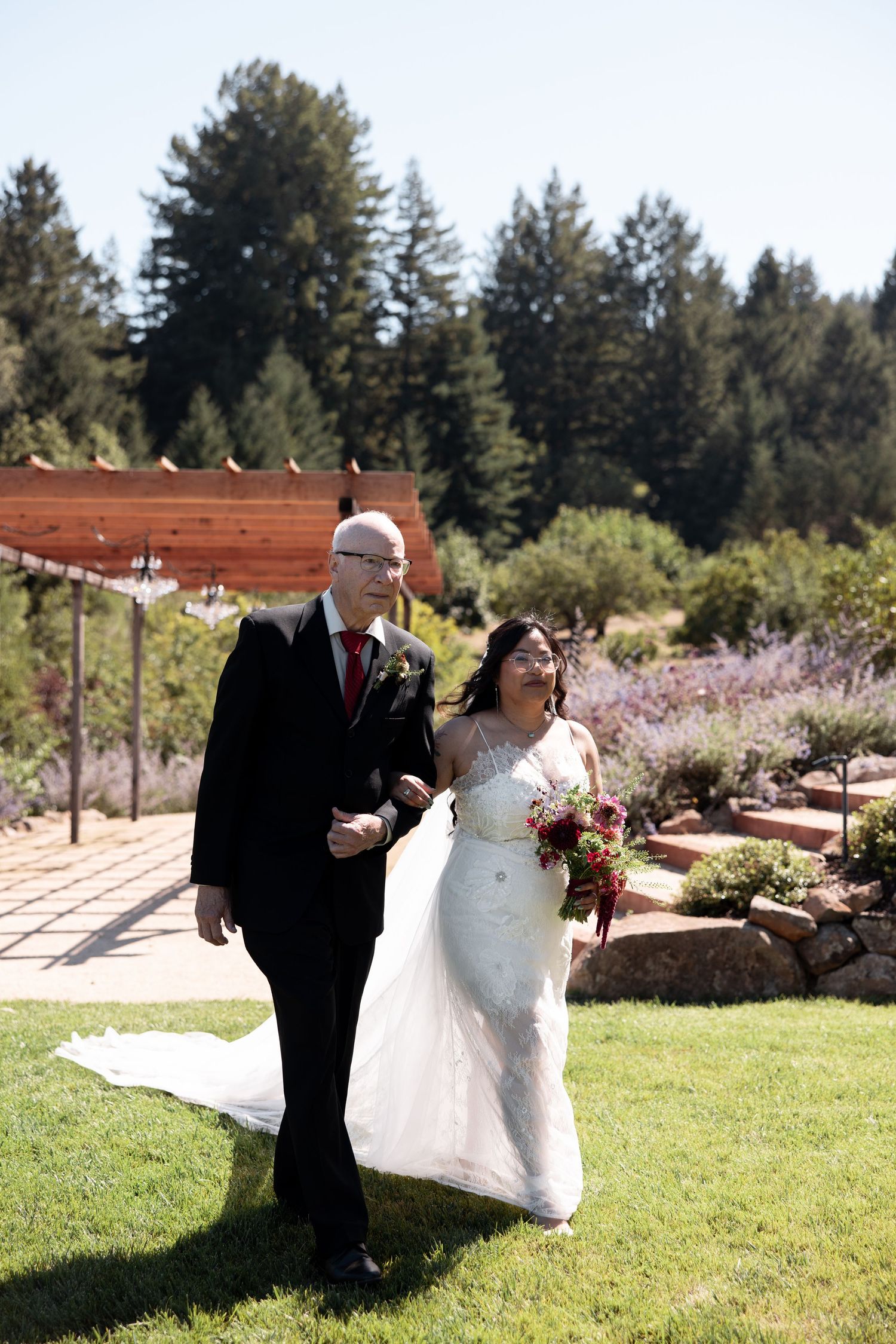 A couple in formal wedding attire walks together on a sunny lawn with evergreen trees in the background.