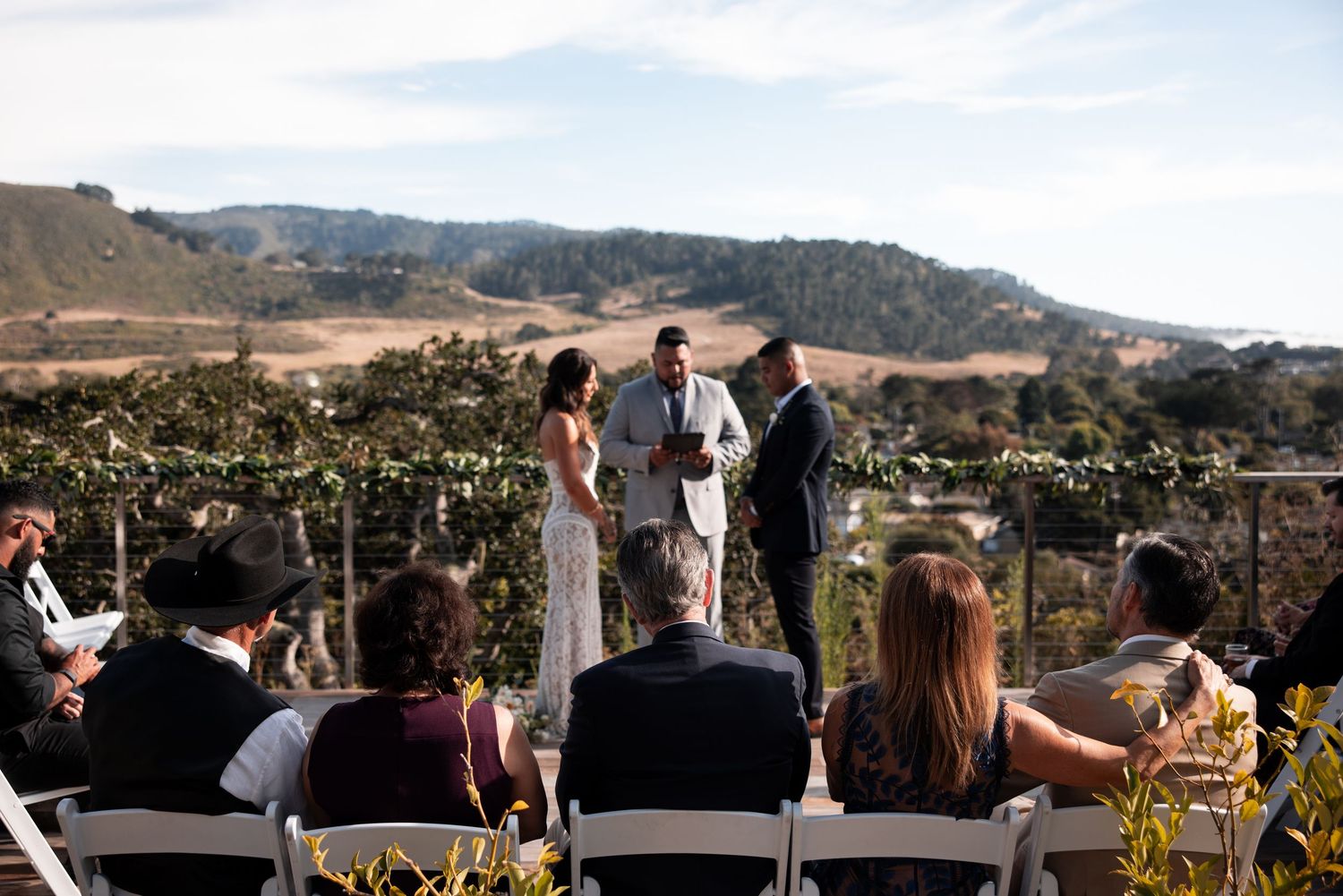 An outdoor wedding ceremony takes place at a vineyard with rolling hills and mountains in the background on a sunny day.
