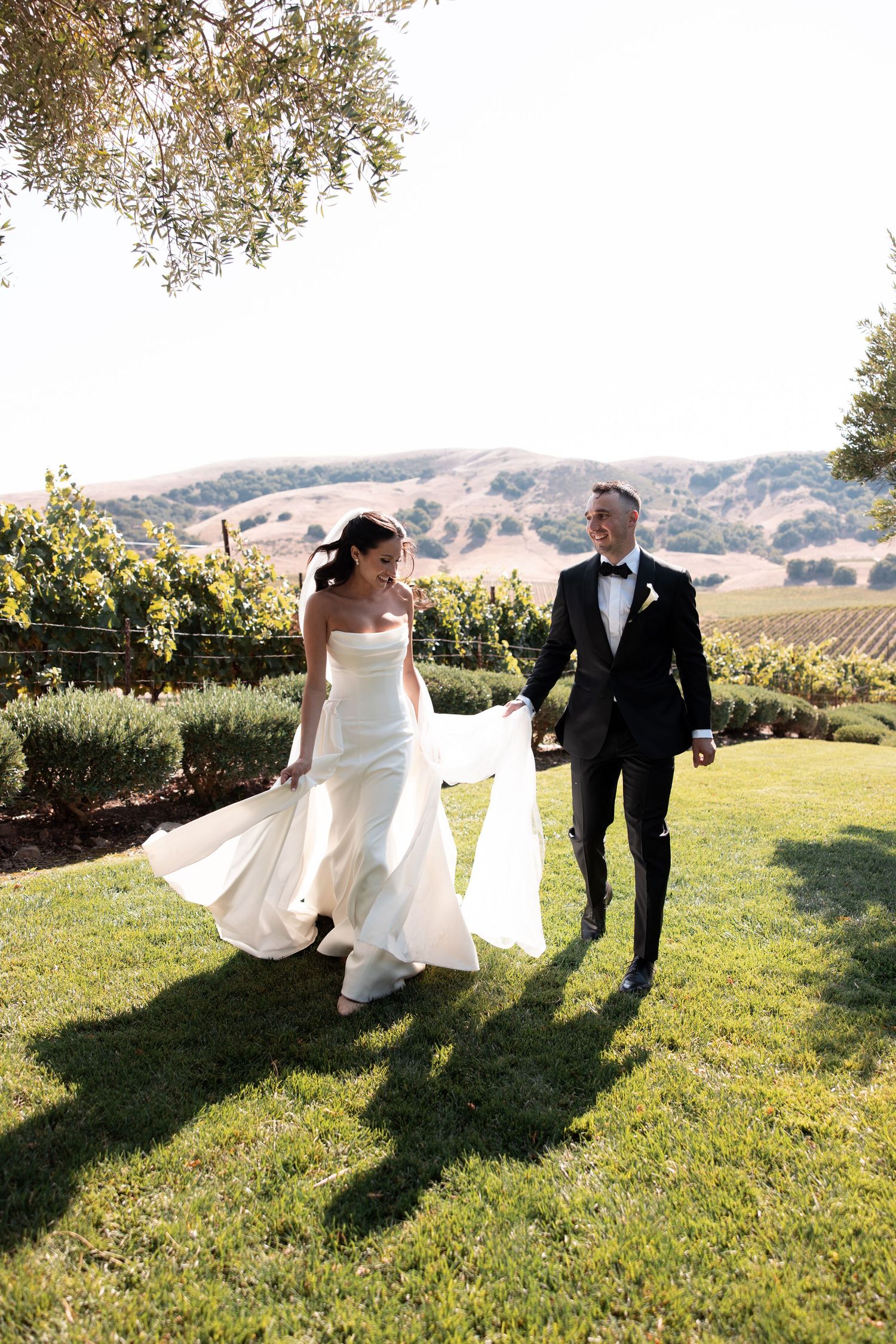 A couple strolls through a scenic vineyard on a sunny day with rolling hills and grapevines in the background.