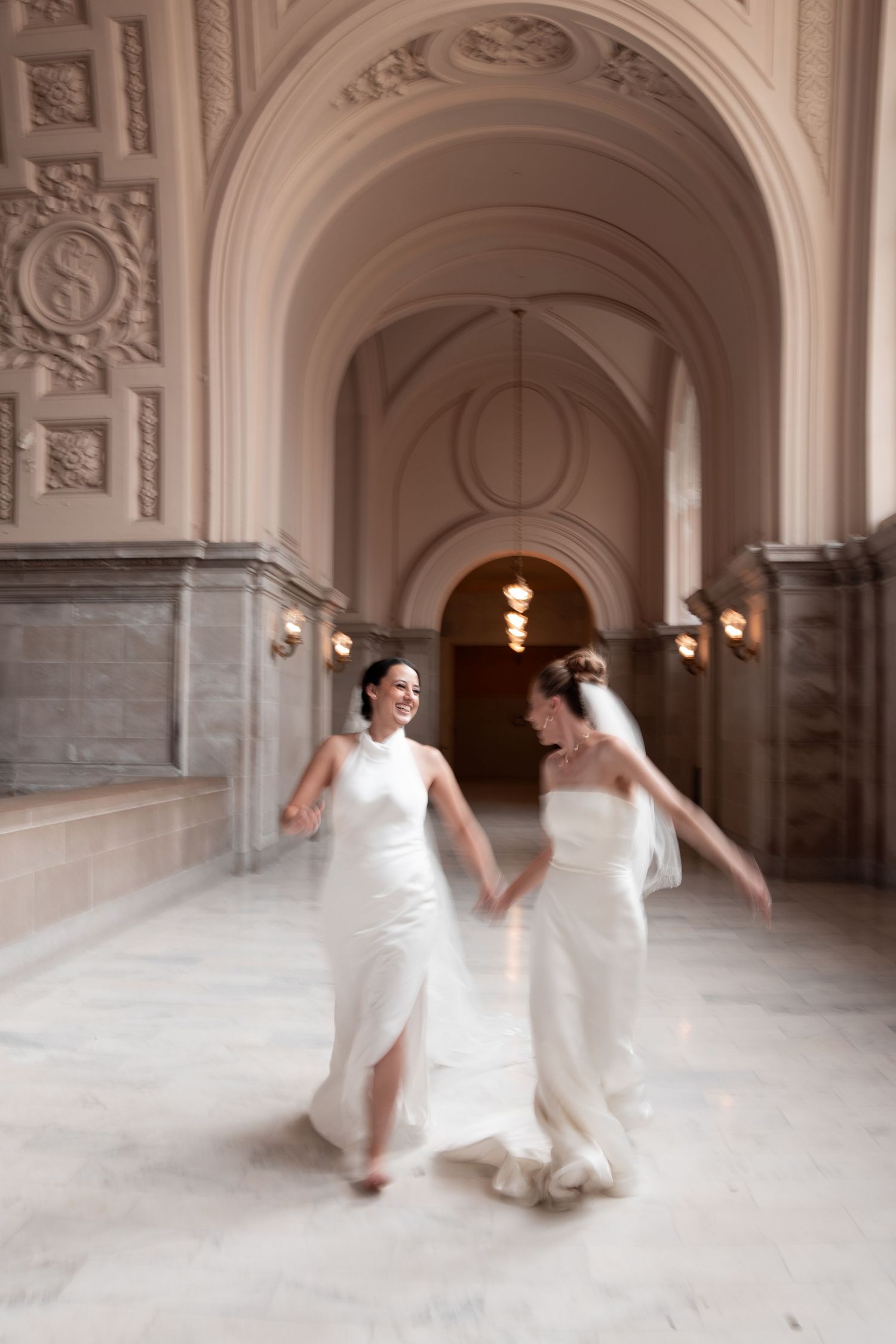 Two brides in white gowns hold hands and dance joyfully in an elegant marble hall with ornate arched ceilings.