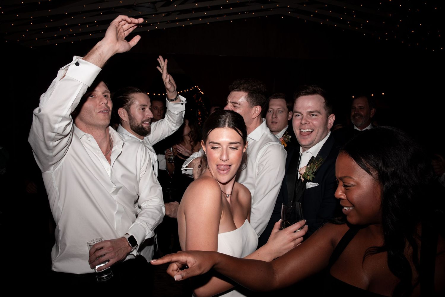 Wedding reception guests dancing joyfully, bride in white dress surrounded by groomsmen in white shirts.