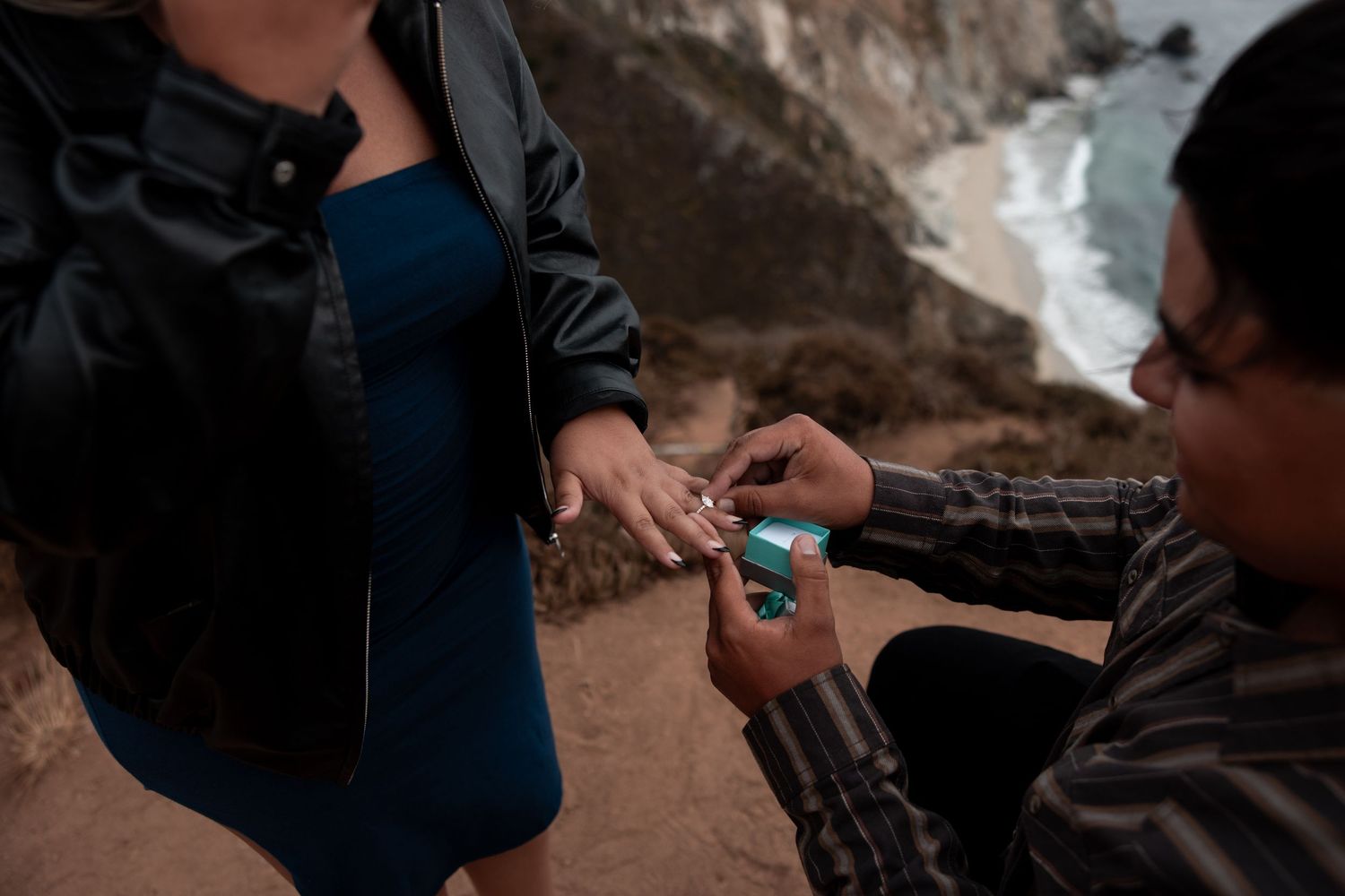 A person in a blue jacket pours water into a small cup while others gather around outdoors in a rocky setting.