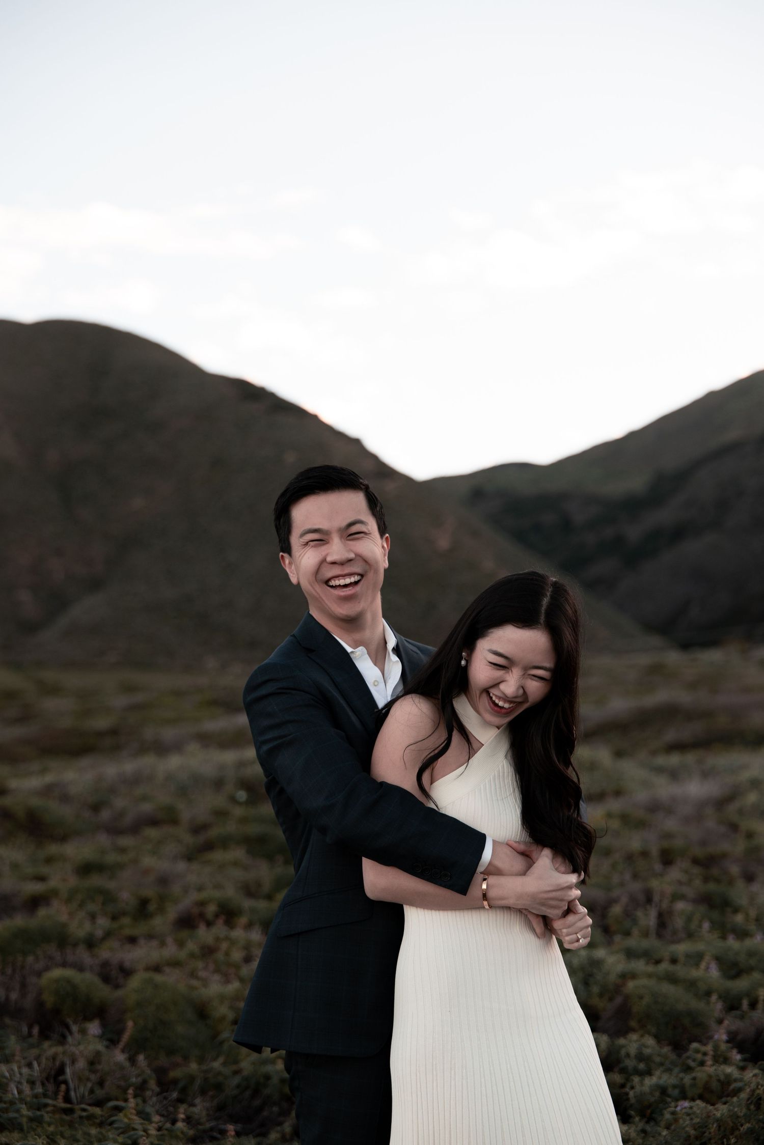 A couple shares joyful laughter together in a scenic mountain landscape during a romantic photoshoot.