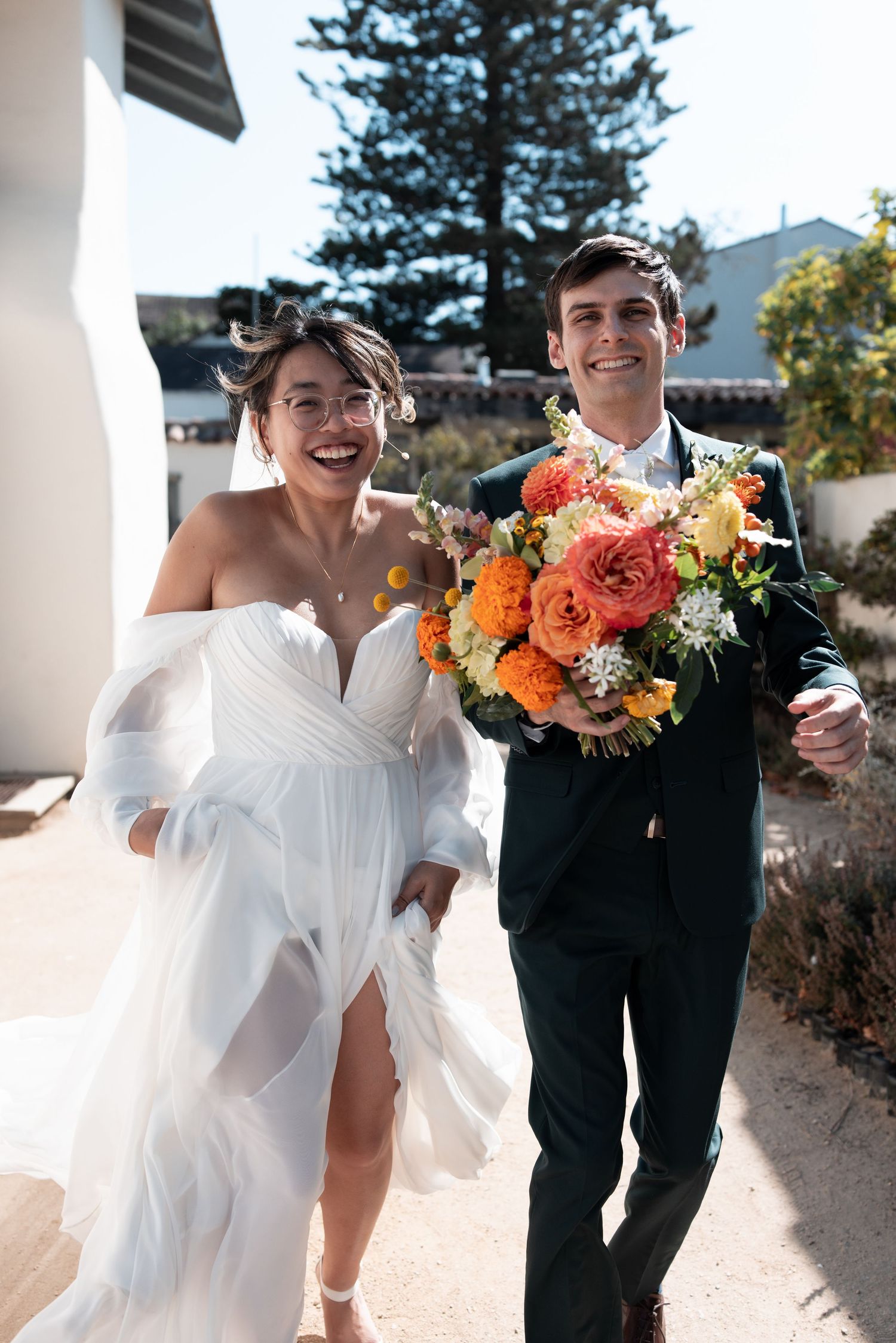 A couple shares joyful moments outdoors in wedding attire with vibrant orange and peach floral bouquets.