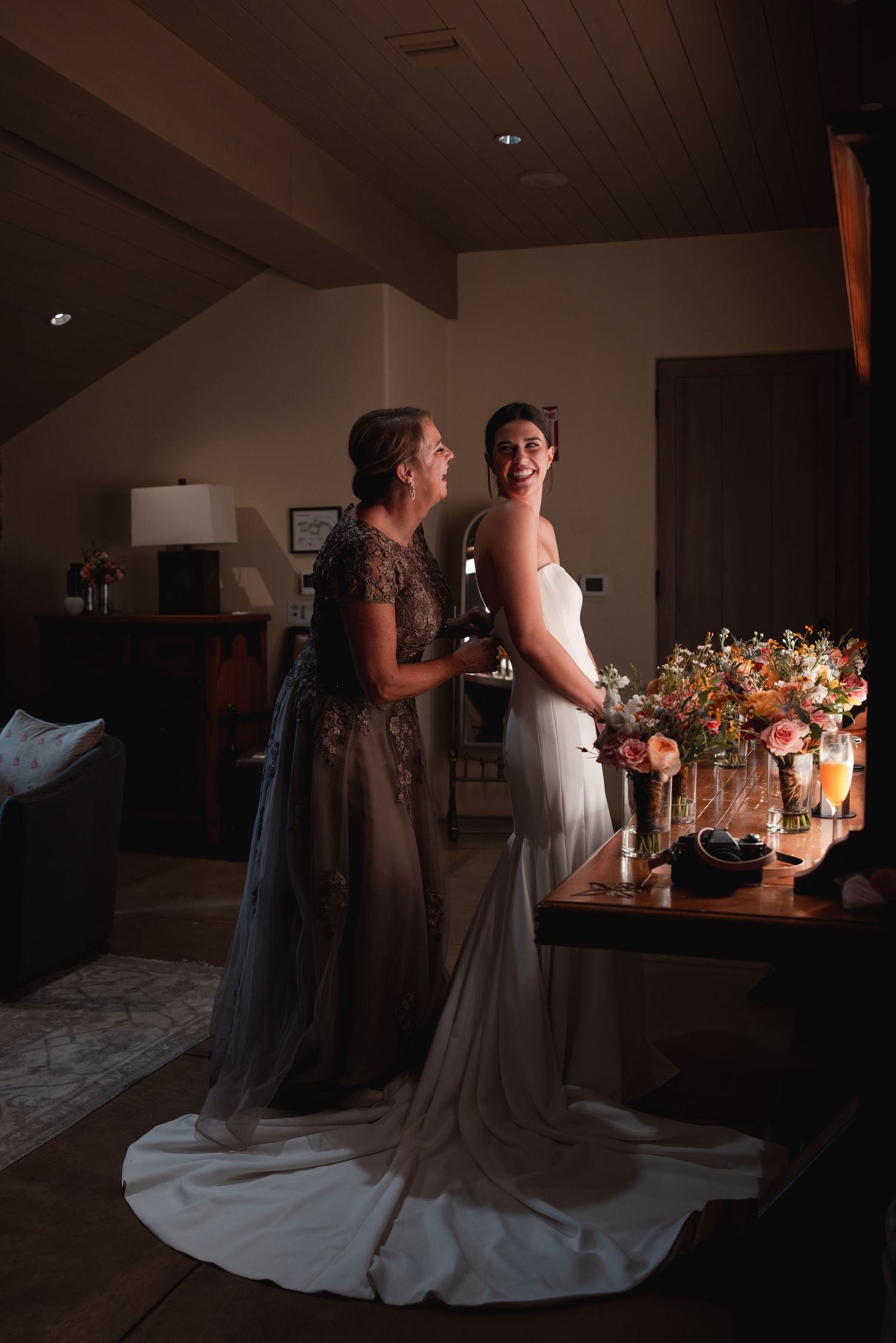 Bride and mother share a joyful moment in a dimly lit room, with a floral arrangement nearby.
