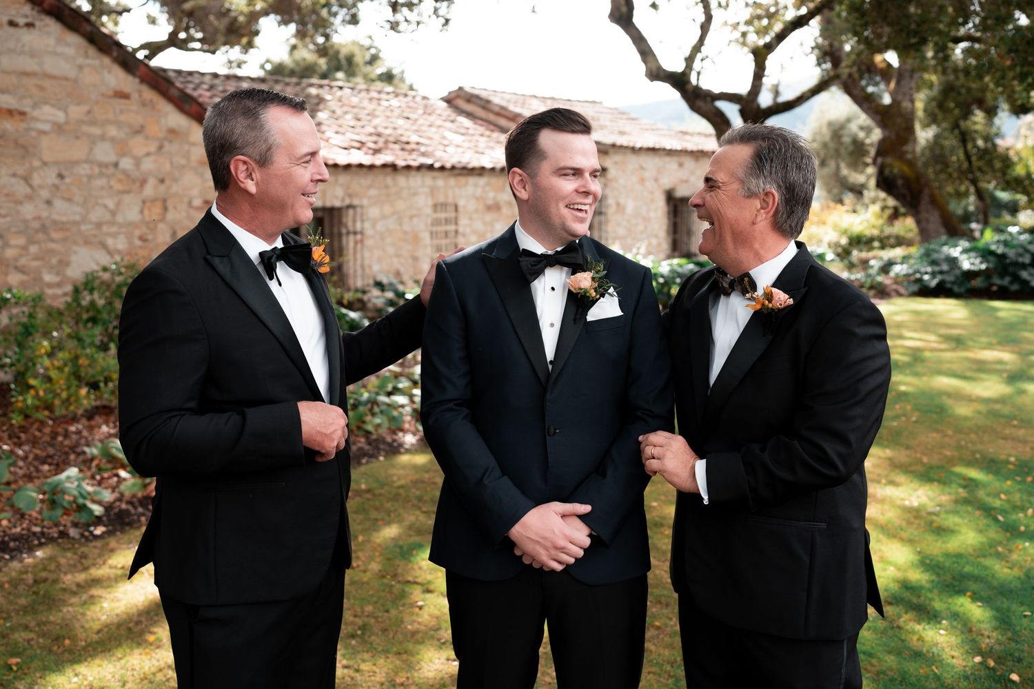 Three men in black tuxedos with bow ties laugh together outdoors at a wedding celebration.