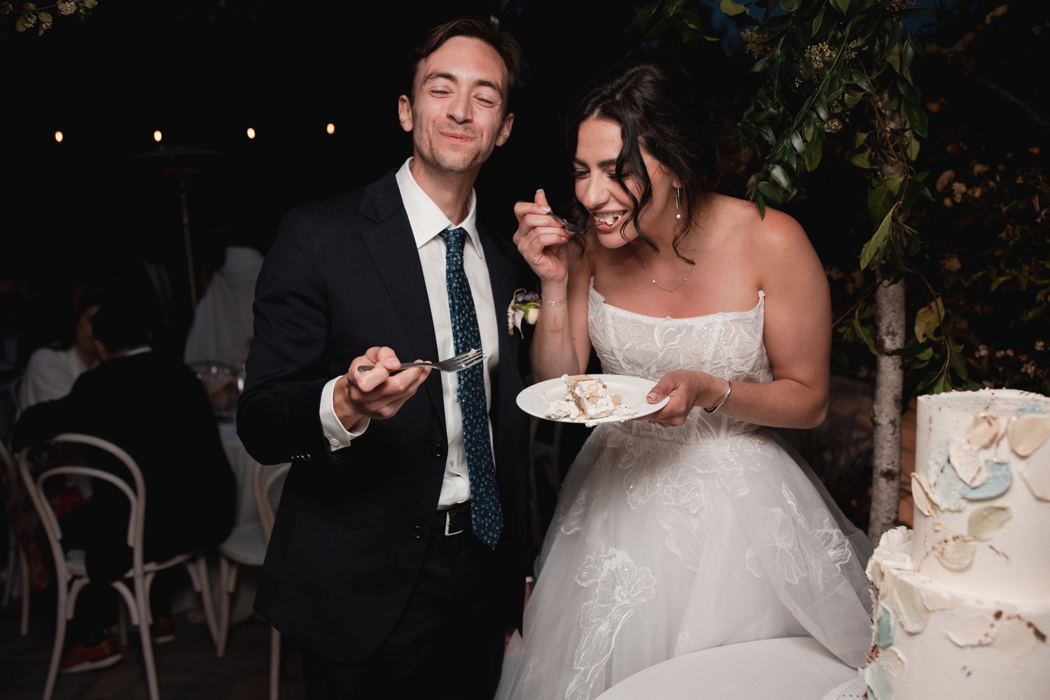A wedding couple cuts into their elegant white tiered cake at an evening reception celebration.