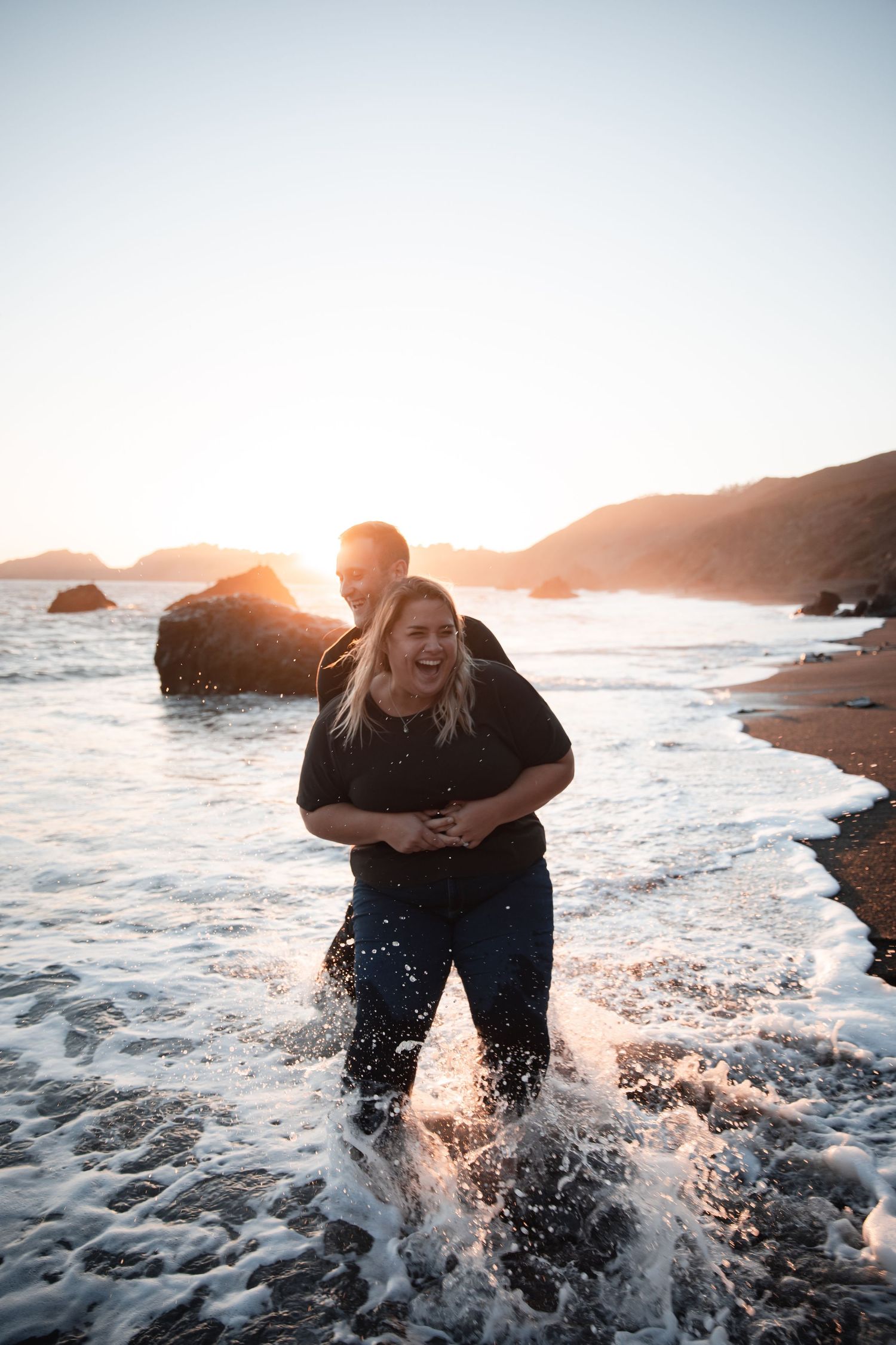 A couple plays in the ocean waves at sunset, splashing water and embracing along a rocky coastline.