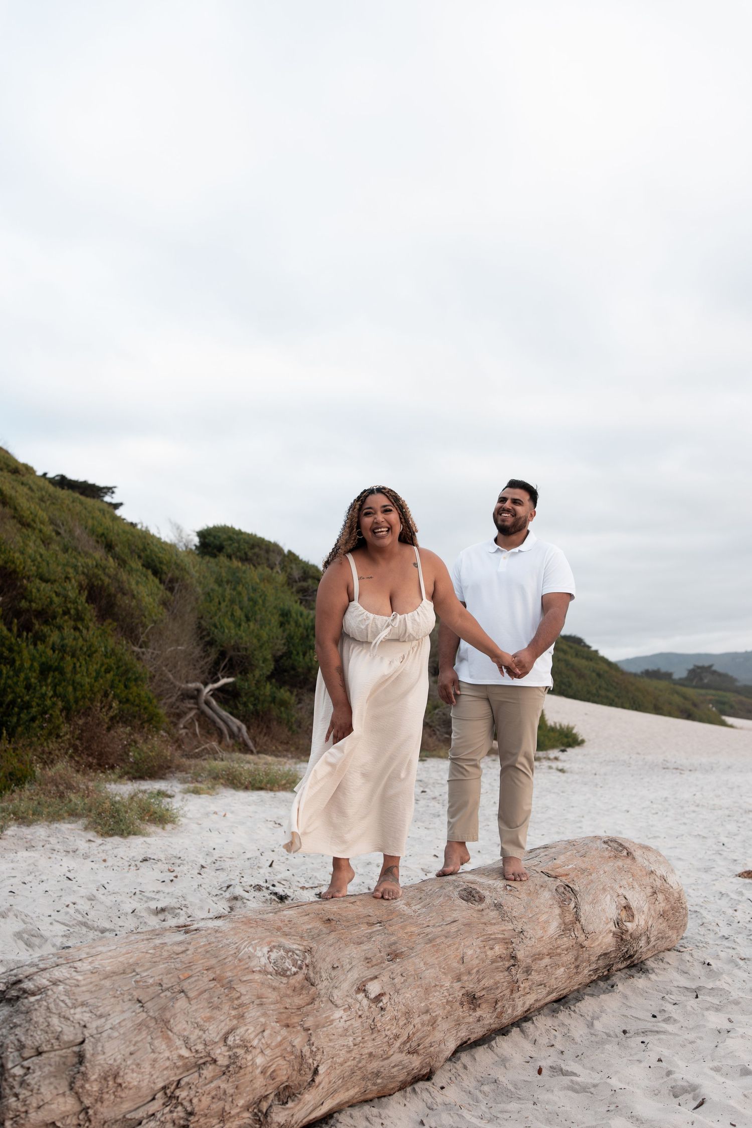 A couple sits together on a large driftwood log at a scenic beach with mountains in the background.