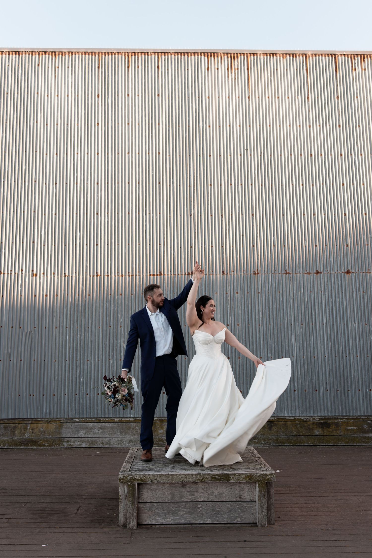 A couple in wedding attire poses playfully on a wooden platform against an industrial metal wall.