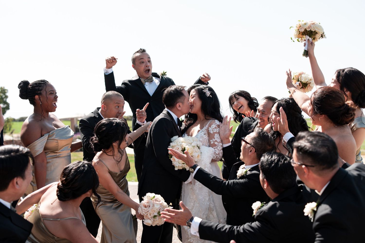 Joyful wedding party celebrating bride and groom kissing outdoors, surrounded by bridesmaids in gold dresses and groomsmen.