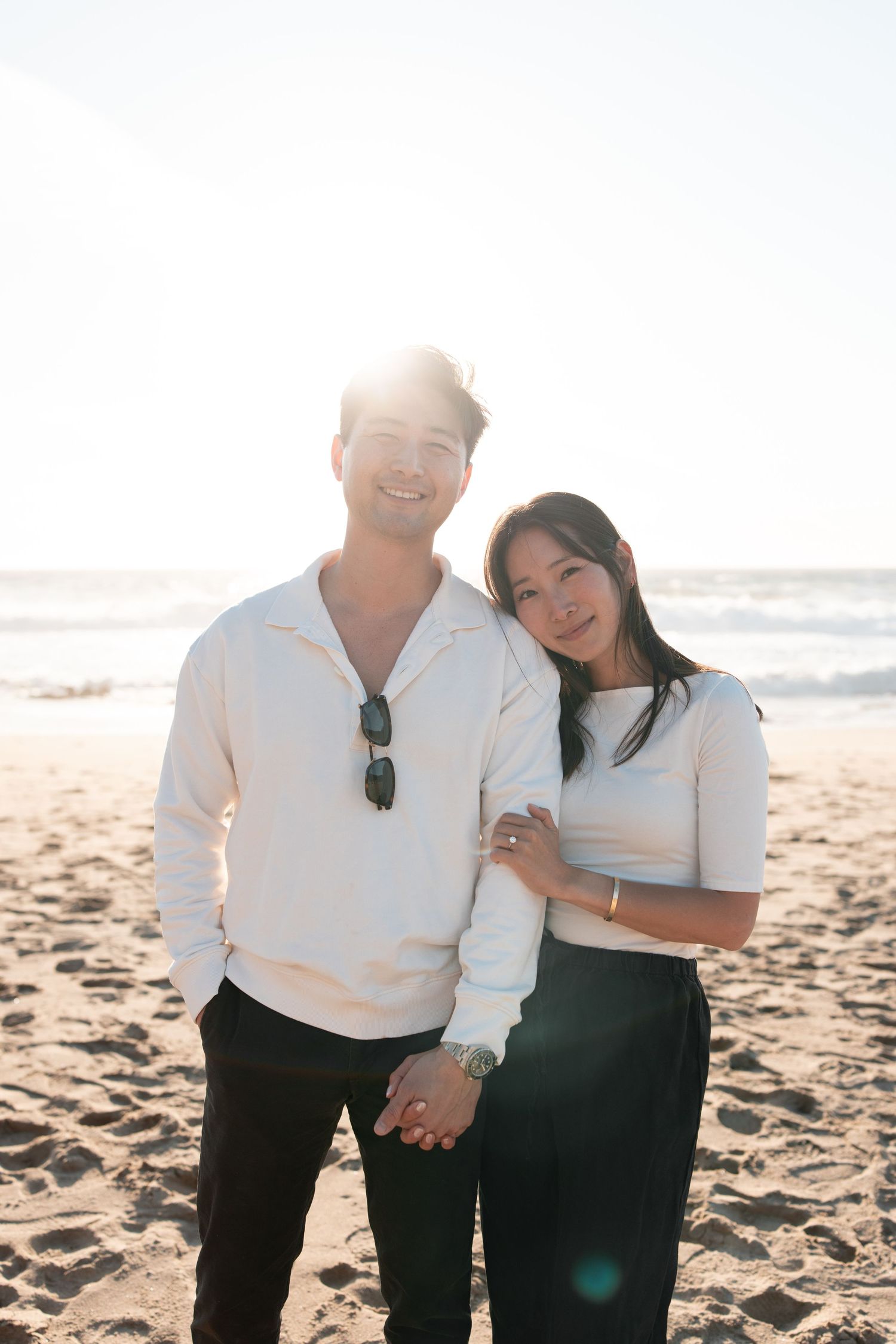 Couple holding hands on a sunny beach, wearing white tops and black pants, smiling with ocean waves in the background.
