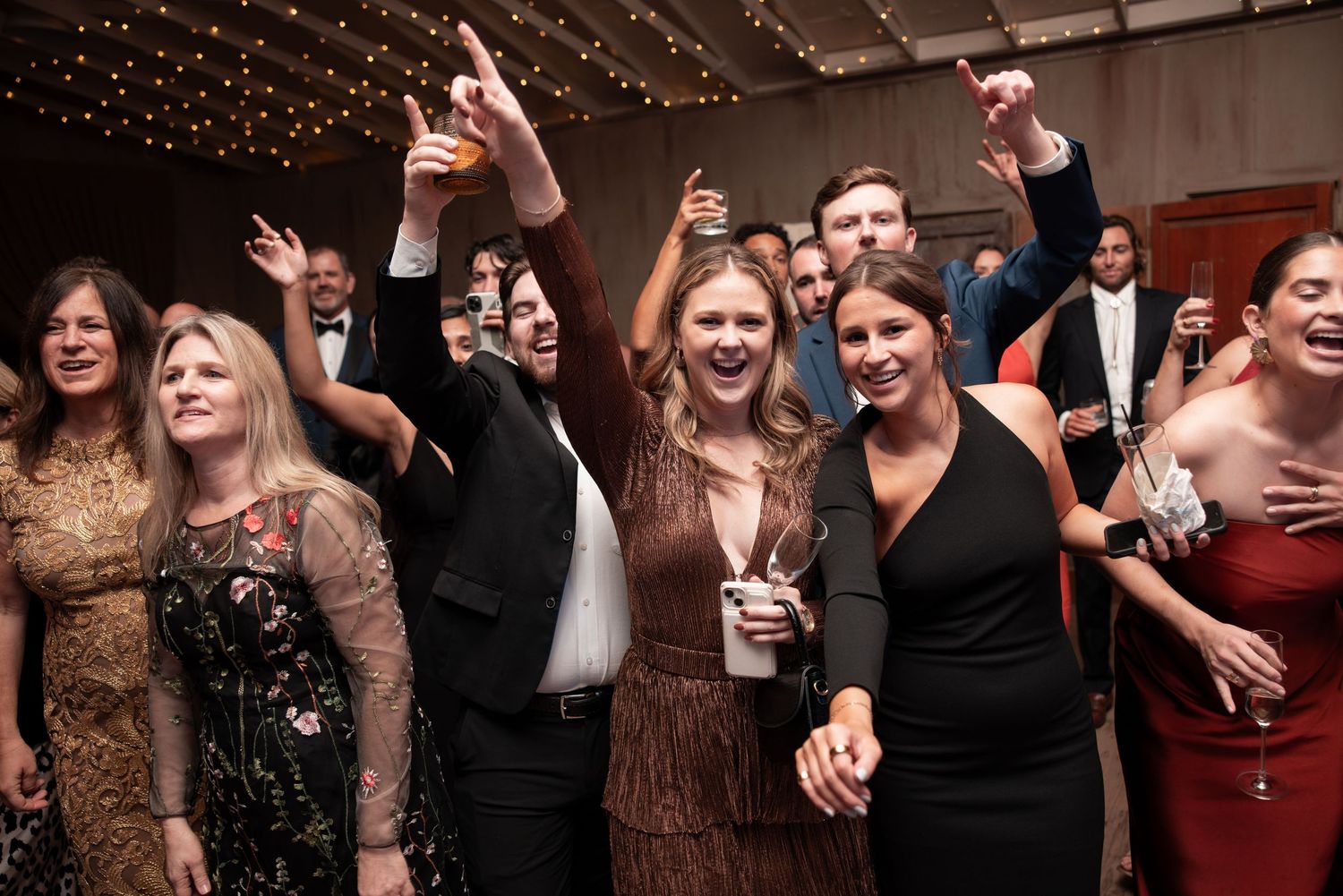 Guests celebrating at a formal party, raising drinks and dancing with joy under warm string lights.