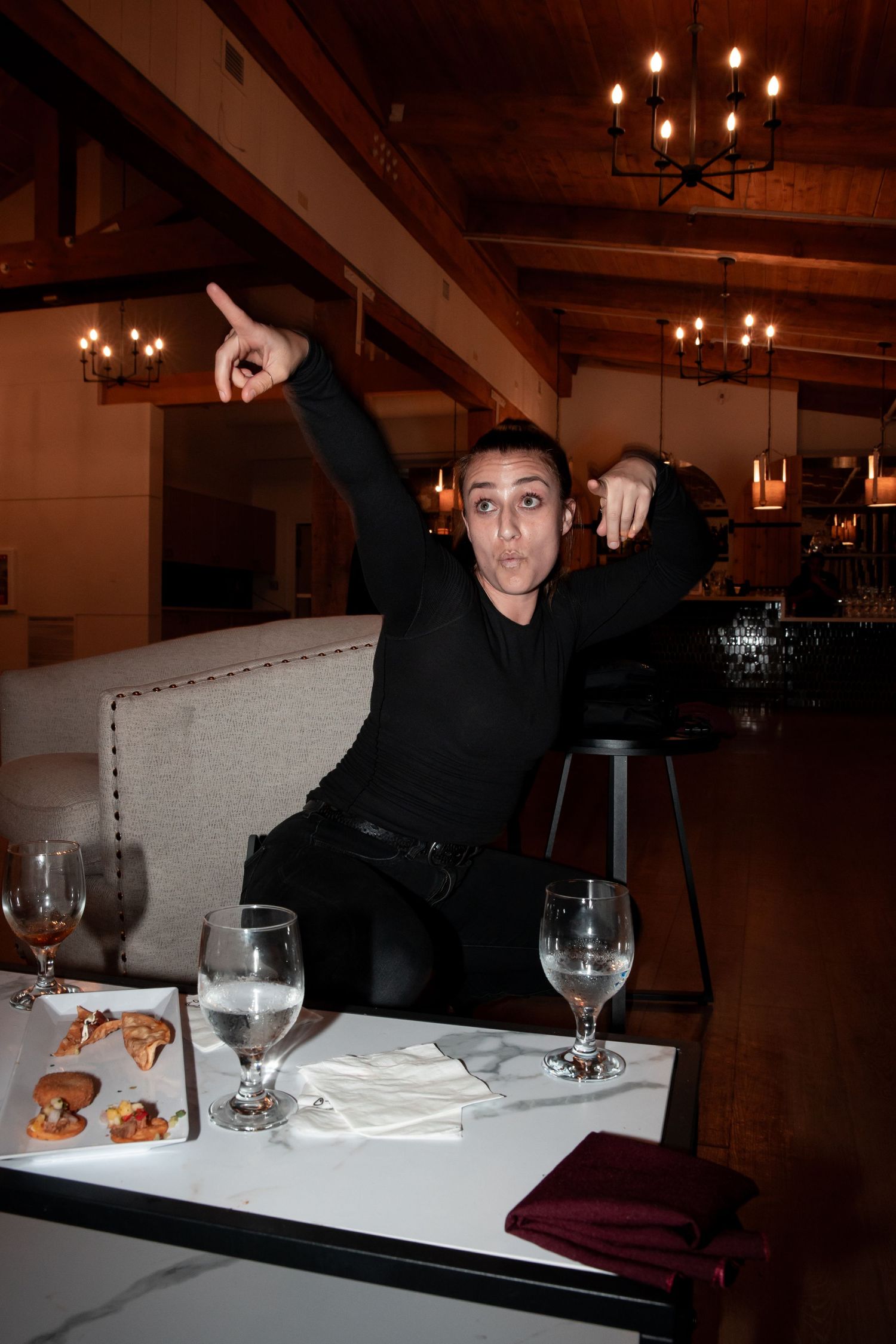Person in all black attire strikes a dramatic pose at a restaurant table with wine glasses and plates.
