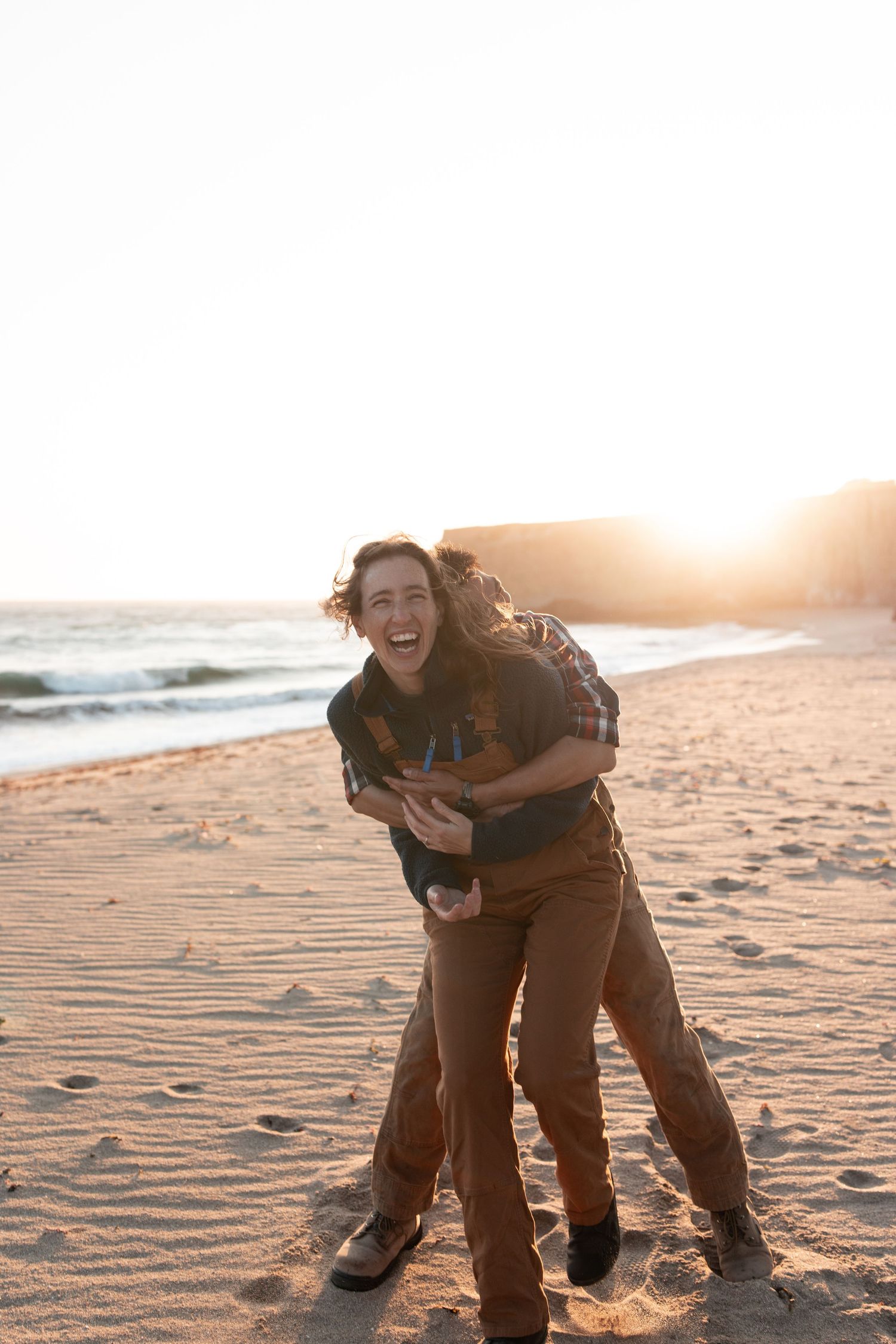 A couple shares joyful moments together on a sandy beach at sunset, creating playful and candid interactions.