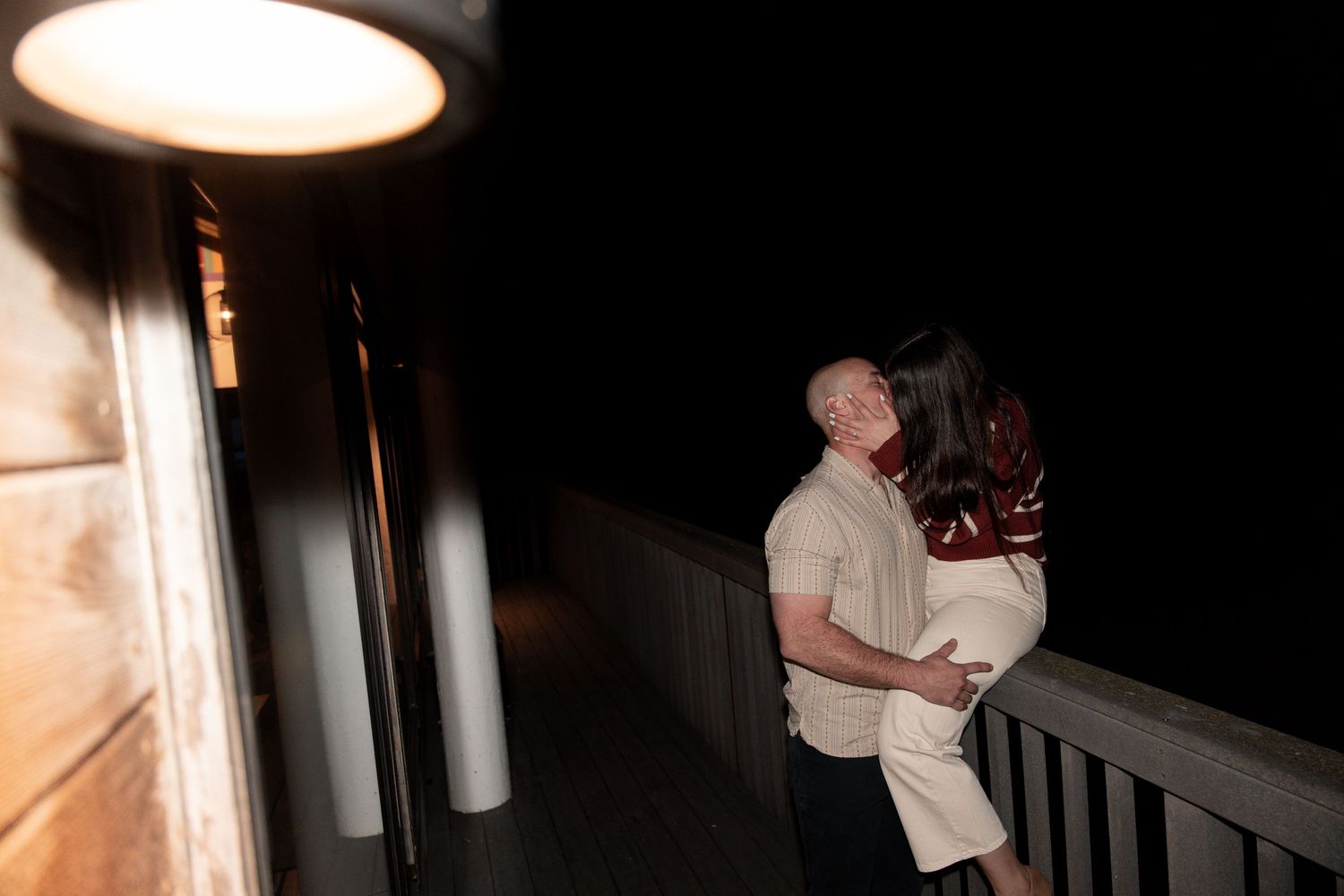 A couple shares an intimate moment on a dark balcony under outdoor lighting at night.