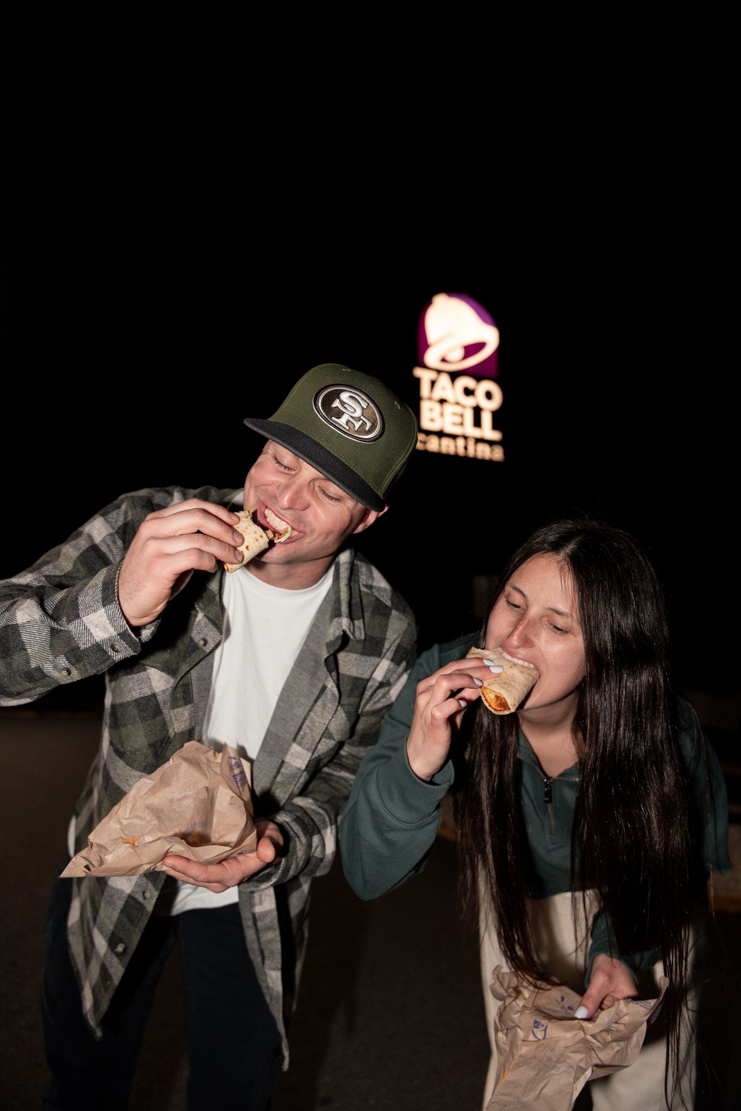 Two people share drinks from bottles at night in a casual outdoor setting.