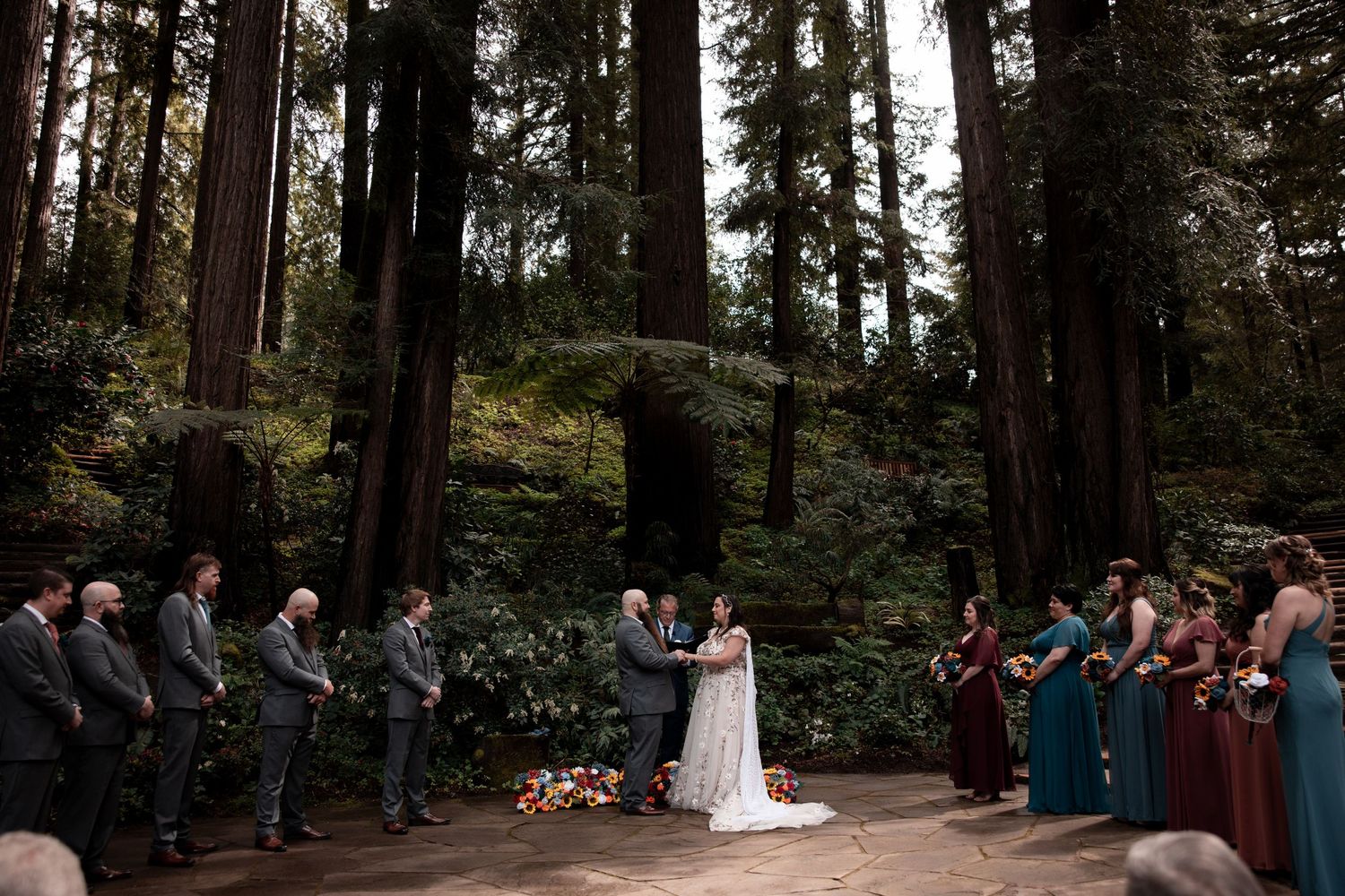 A wedding ceremony takes place in a serene forest setting surrounded by tall evergreen trees and gathered guests.