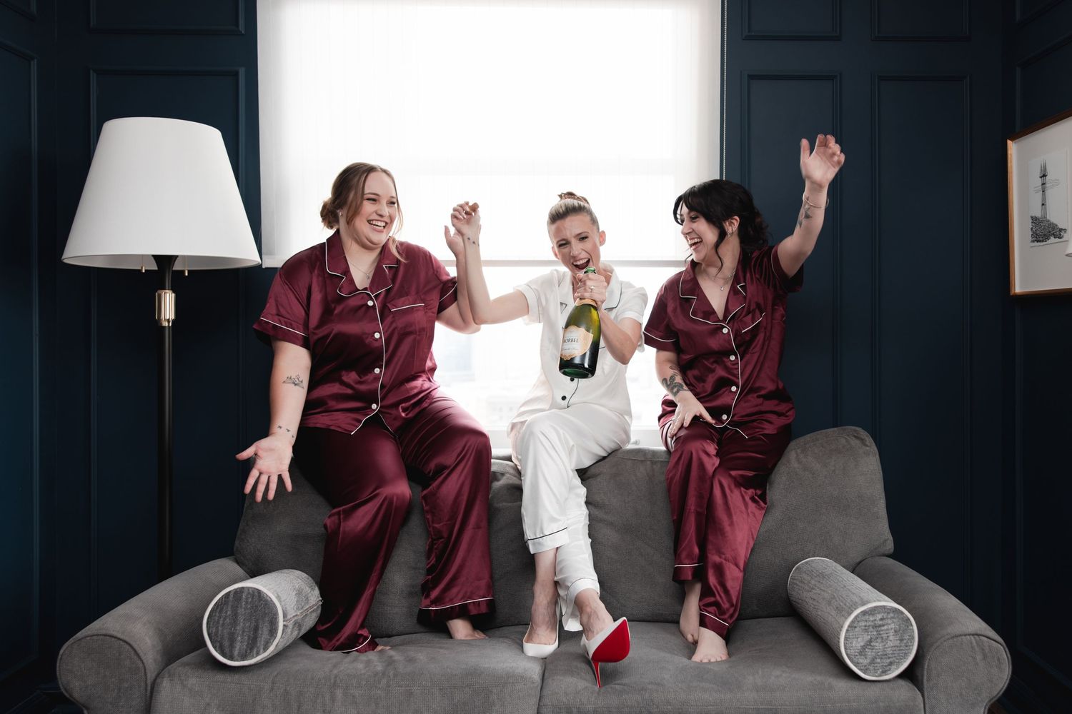 Three women in satin pajamas celebrate on a couch, one holding a champagne bottle, arms raised joyfully.