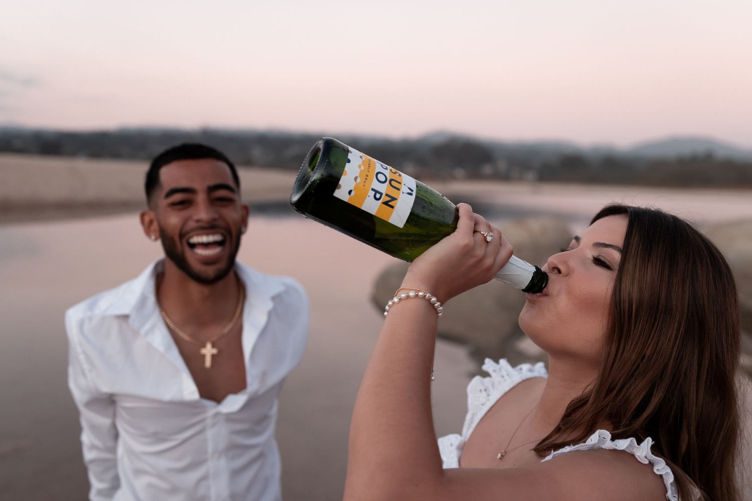 A couple shares a romantic moment while drinking champagne on a beach at sunset.