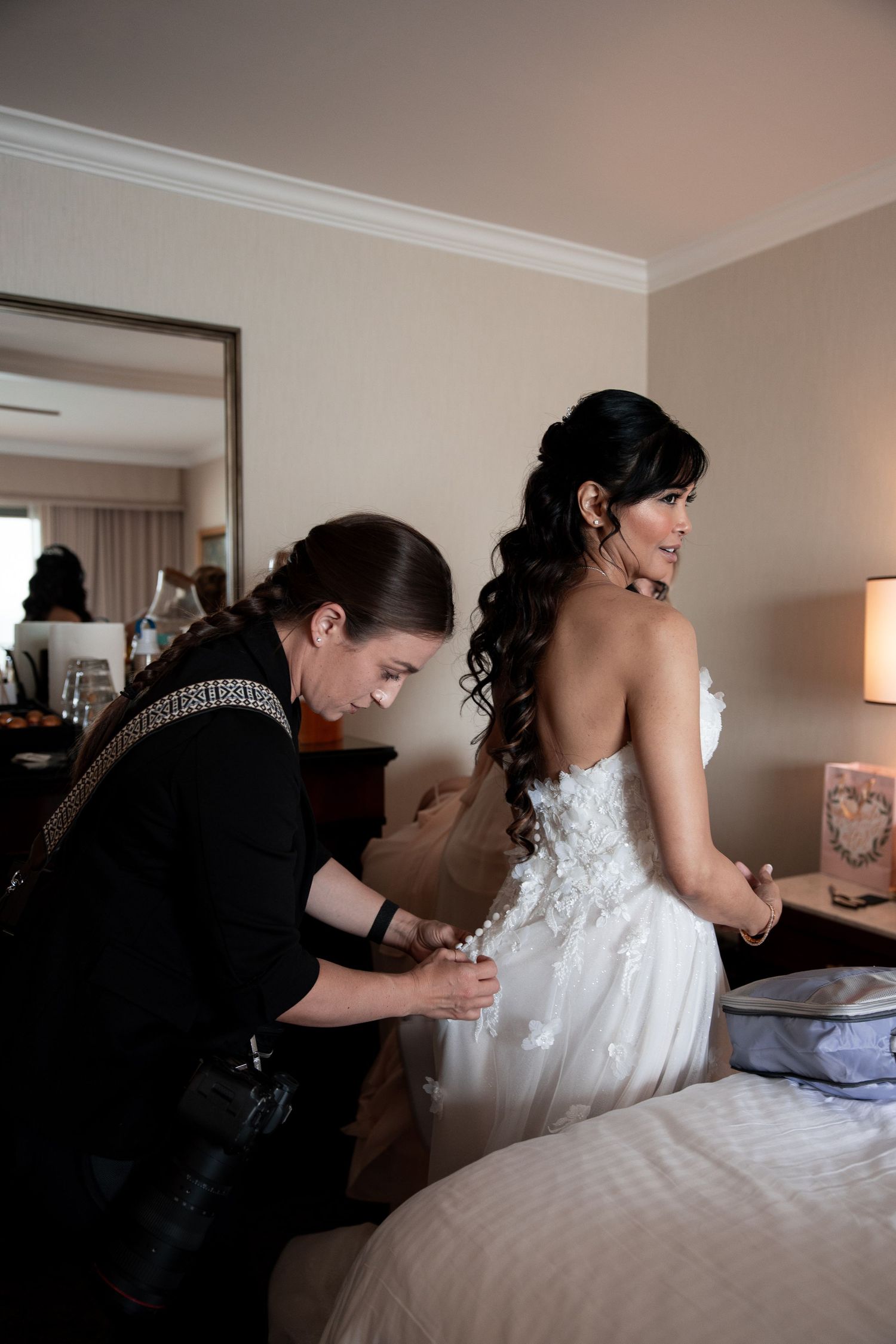A bridesmaid helps a bride in a white lace strapless gown with her dress in a hotel room.