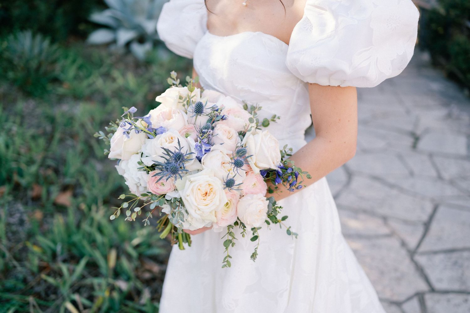 Elegant bridal bouquet with white and pale blue flowers held against a white wedding dress with puffy sleeves.
