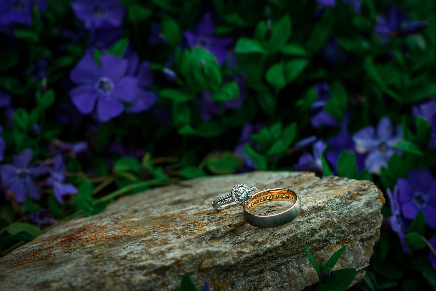 Wedding rings displayed on rustic wood with purple flowers in the background.
