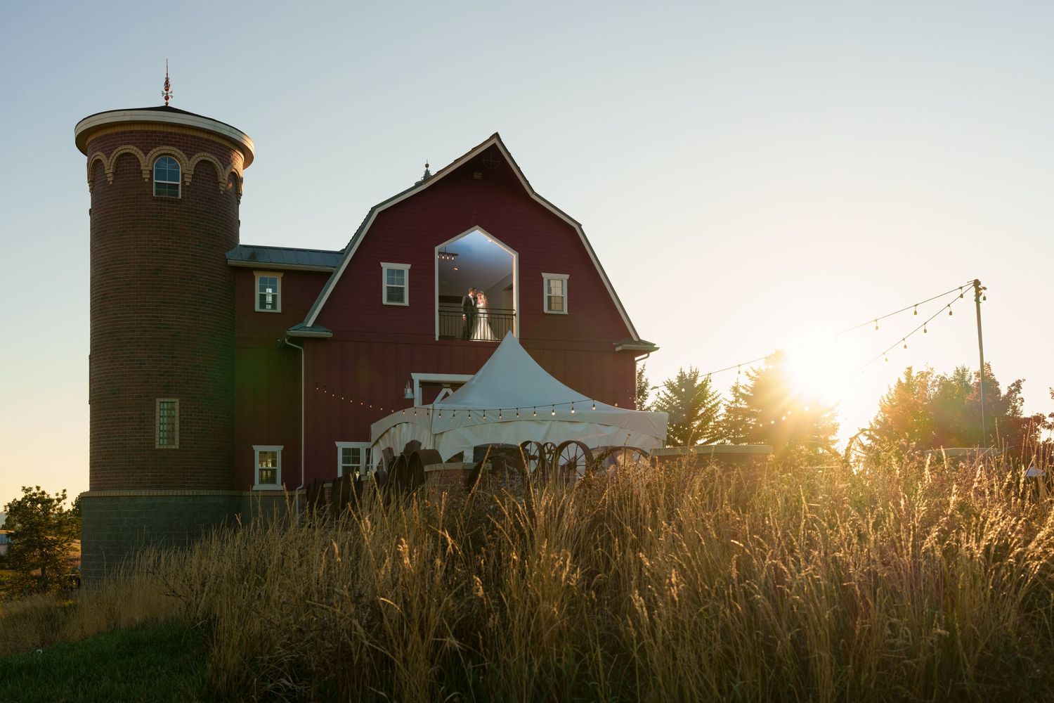 Red barn wedding venue at sunset with rustic silo tower and golden wheat field in foreground.