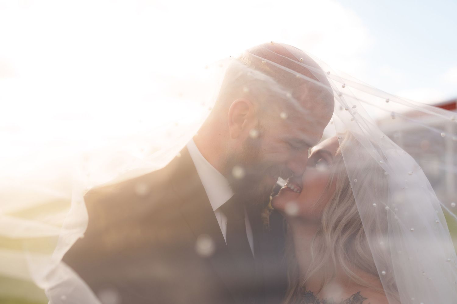 Sun-flared intimate moment between newlyweds through flowing veil.