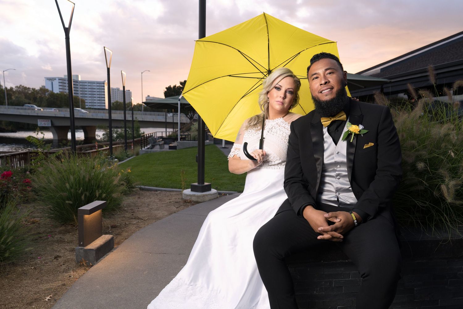 A wedding couple sits outdoors holding a bright yellow umbrella at dusk.
