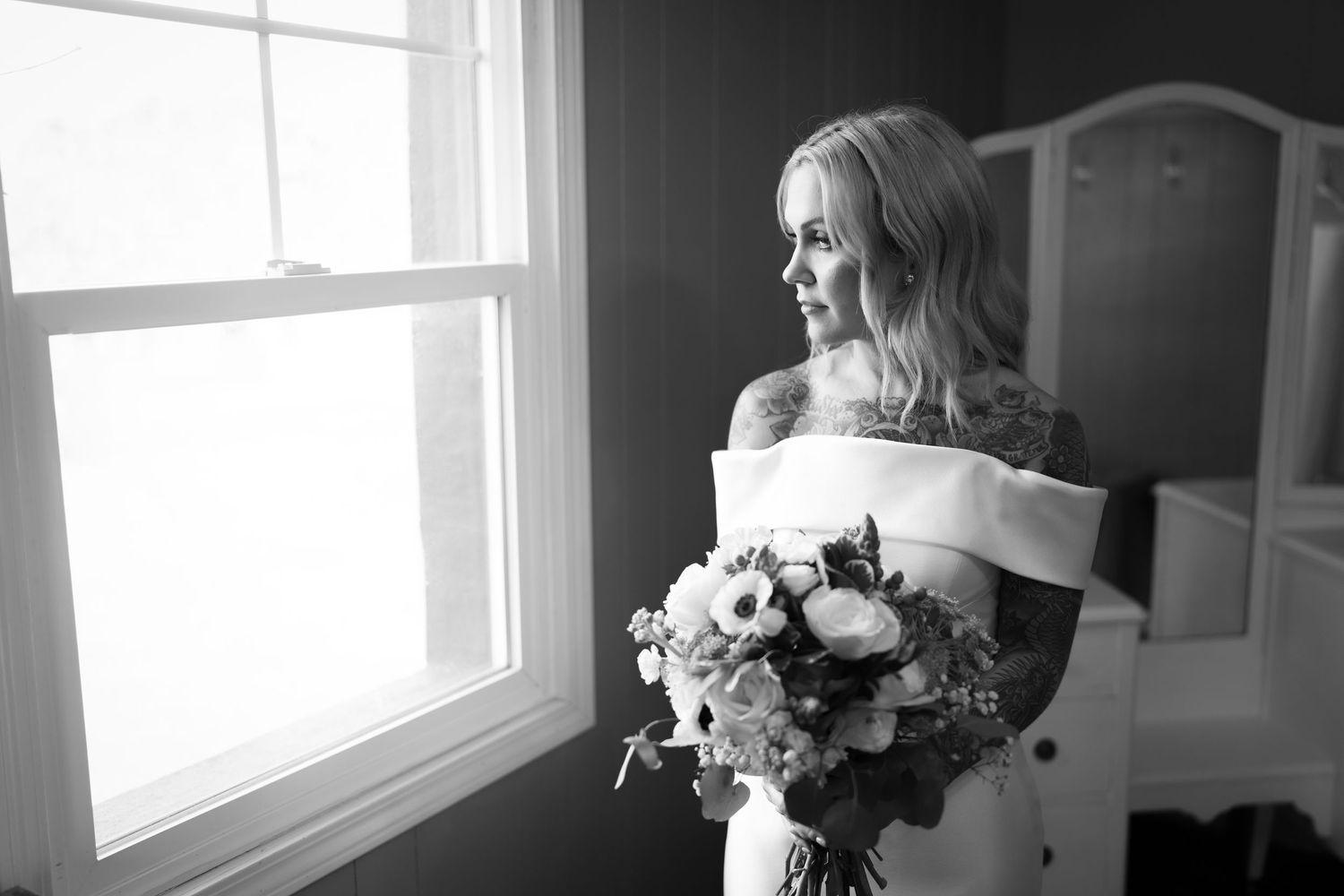 Black and white portrait by window showing figure holding elegant floral bouquet.