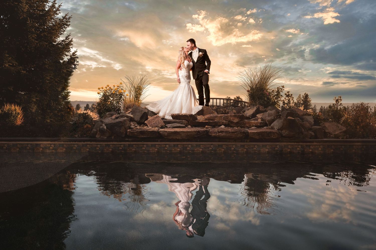Couple standing on rocks with dramatic sunset clouds reflected in still water below.