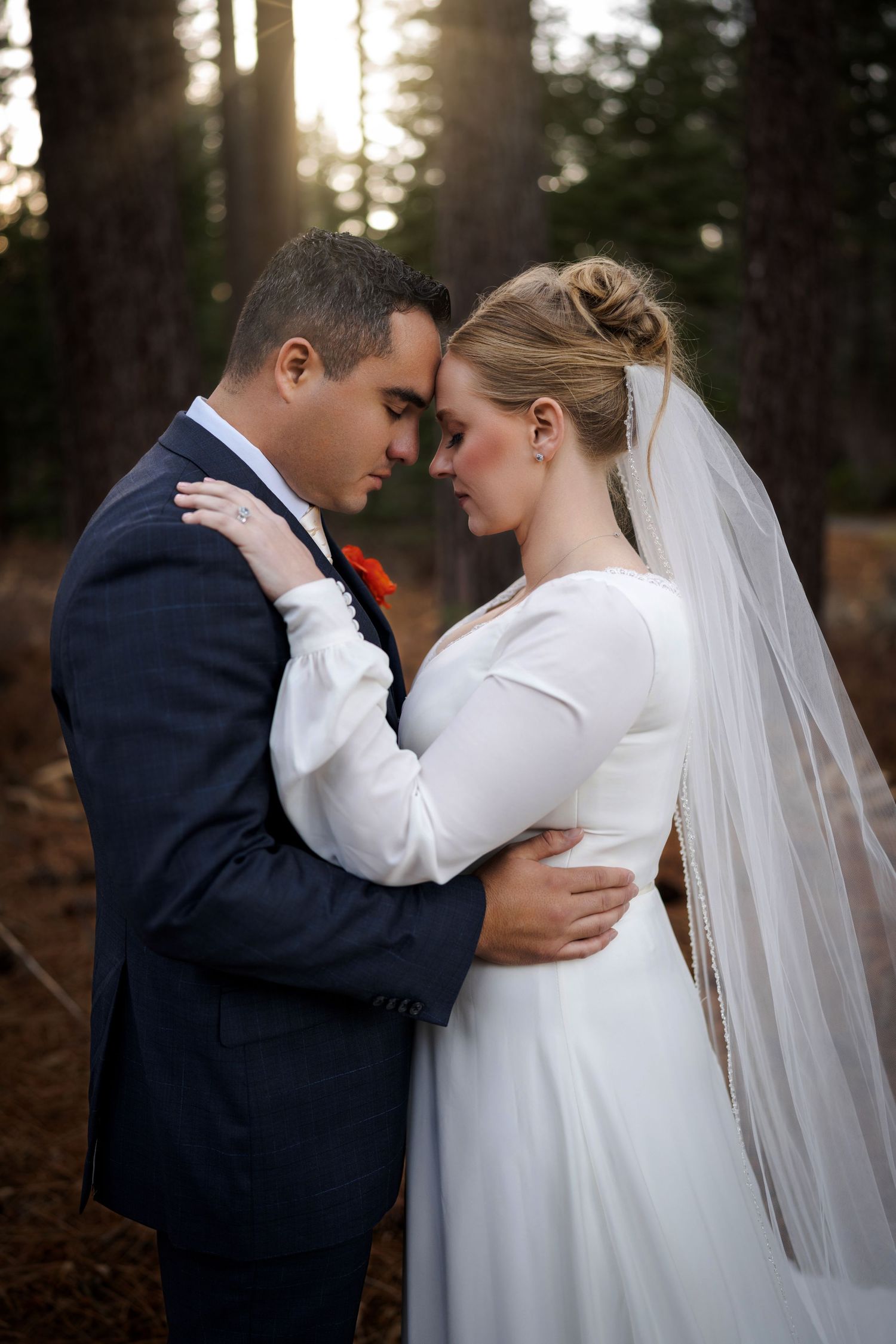 Intimate forest wedding portrait of newlyweds embracing among tall pine trees at sunset.